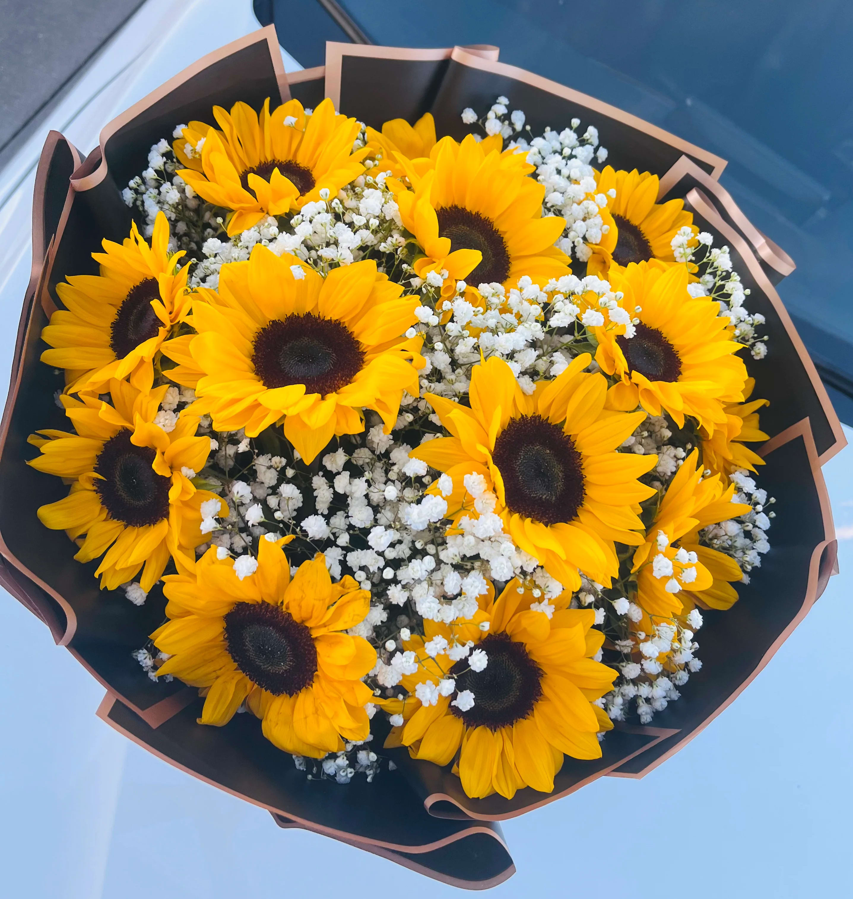 Sunflower Shine - A ring of bold yellow sunflowers with dark centers creates an immediate sunny impression-perfect as a birthday bouquet, a Father's Day gift, or a cheerful "thinking of you" arrangement. Sunflower Shine is a hand-tied flower arrangement that sends a message of warmth and celebration, whether you're congratulating a new graduate, brightening a get-well day, or saying thanks to someone who made your season better.  The bouquet pairs tightly gathered sunflowers with clusters of small white filler blooms that weave through the heads for soft contrast and added texture. The stems are finished in decorative dark wrapping paper with a tan edge for a casual yet polished presentation that suits housewarmings, retirements, or simple everyday surprises. Size and stem count are kept compact for easy display; treat with fresh water and recut stems on arrival to prolong vase life.  Sunflower Shine works year-round-its sunny palette lifts spirits for spring and summer events and brings a fresh note to holiday gatherings like Thanksgiving or Christmas when you want something bright and unexpected. Order for birthdays, graduations, Mother's Day, or "just because" moments when you want the recipient to feel genuinely seen and celebrated.