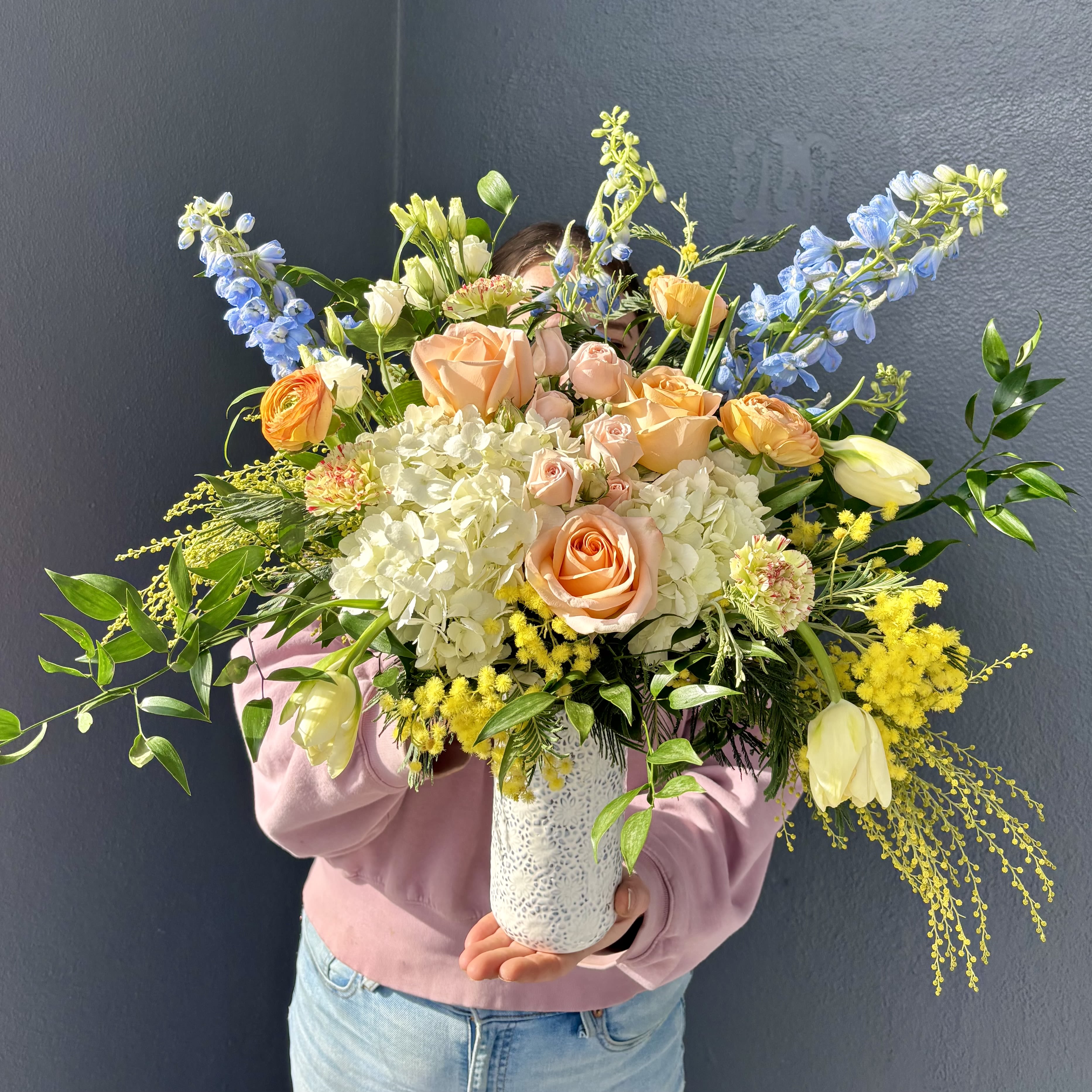 Walking on Sunshine - A bright and cheery seasonal arrangement. Vibrant and fragrant Mimosa compliment calming blue delphiniums and peach roses to make the perfect arrangement for any occasion. This design is only available for the short Mimosa season. Mimosa flowers symbolize strength, sensibility, and solidarity, especially on International Women's Day, March 8th. Its delicate, sunny-yellow blooms and ability to thrive in harsh conditions make it a powerful symbol for perseverance, hope, and the arrival of spring.