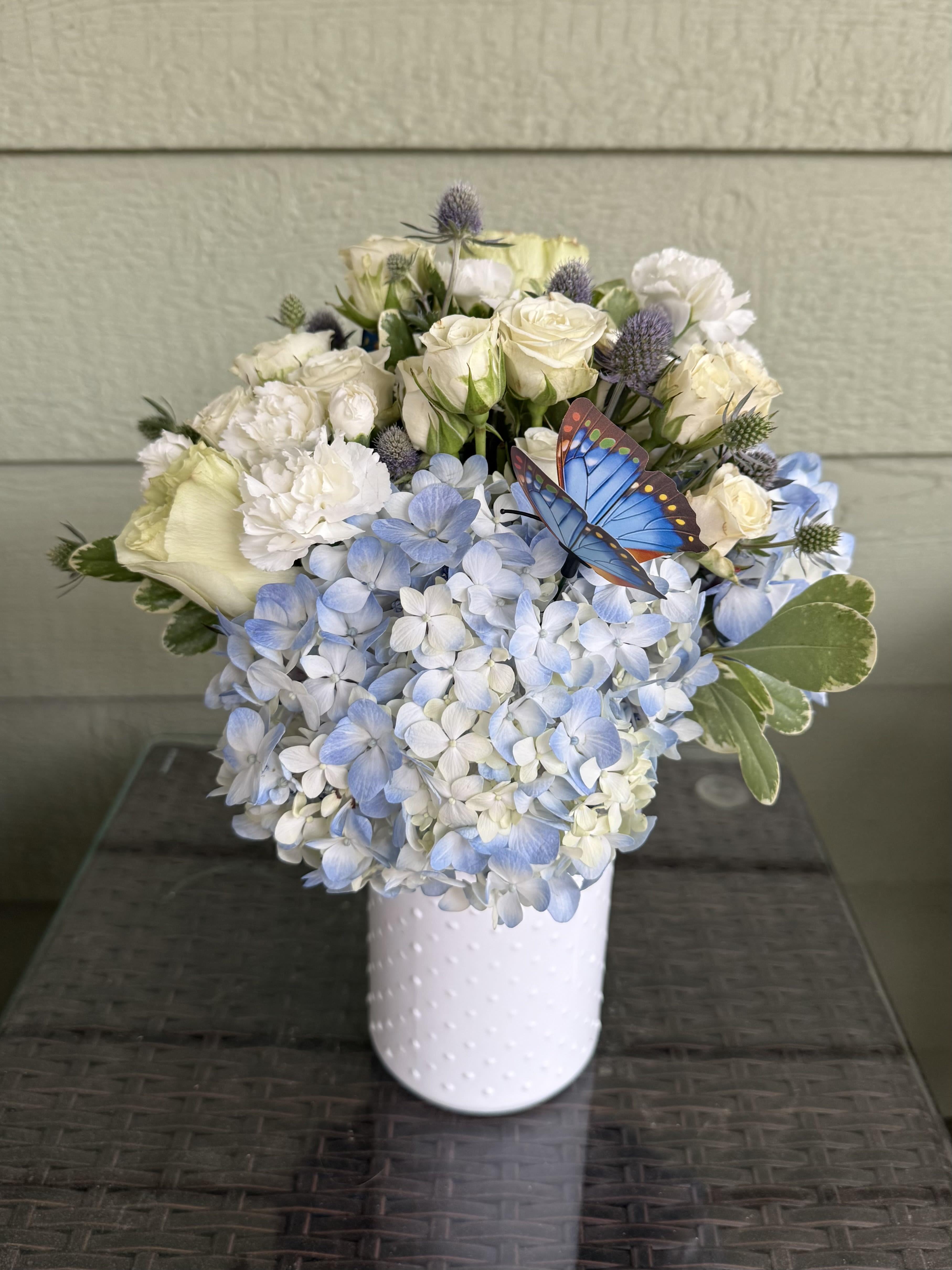 White roses and blue hydrangeas in a white vase with a butterfly accent