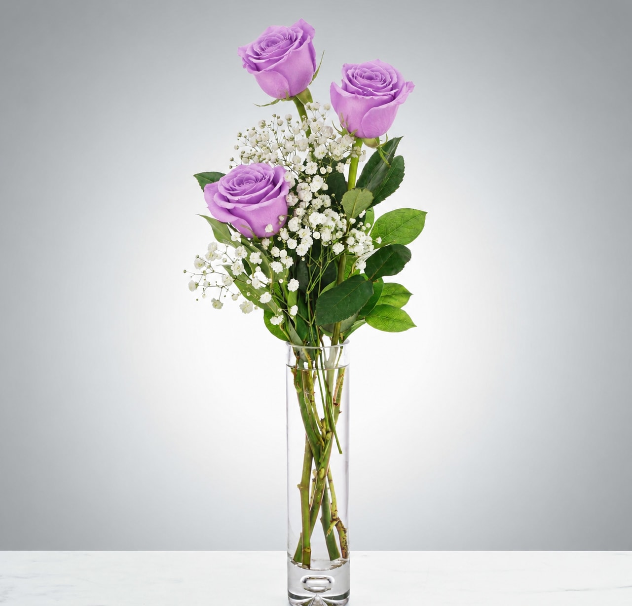 XO Lavander  - Lavender roses rise above a slim clear glass vase in a simple, elegant arrangement made for Mother's Day, birthdays, anniversaries, or just to say "I'm thinking of you." This tall, vertical bouquet-three lavender roses with airy clusters of small white filler flowers and touches of green-creates a calm, romantic silhouette that reads as both thoughtful and refined for the person you want to celebrate.  The composition is intentionally understated rather than dense: blooms are spaced above the vase for an airy profile that highlights the lavender color and delicate white accents. Arrives in a narrow clear glass vase that showcases the stems and keeps the design light; the arrangement is compact enough for a bedside table, desk, or mantel. Recommended for anyone who appreciates a quiet, elegant expression of affection-no fuss, just a graceful, long-lasting reminder you care.