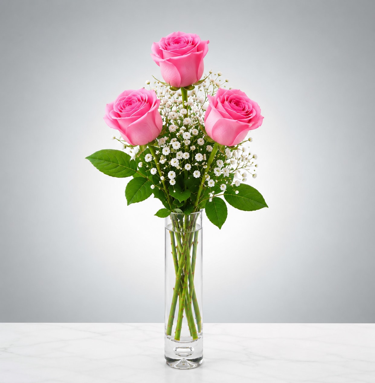 Xo Pink - Three bright pink roses take center stage in this elegant flower arrangement designed for birthdays, romantic gestures, or a cheerful everyday gift. The upright, symmetrical silhouette - one rose perched above two lower blooms - creates a refined, modern look that reads as both joyful and affectionate to the person you're thinking of.  Set in a tall, narrow clear glass vase, the vivid pink petals pop against clusters of small white filler flowers and the visible green stems, giving a fresh, polished presentation. Compact and easy to place, the arrangement arrives ready to display; refresh water every 2-3 days, trim stems at an angle if needed, and it will continue to brighten a bedside table, desk, or kitchen counter for several days.