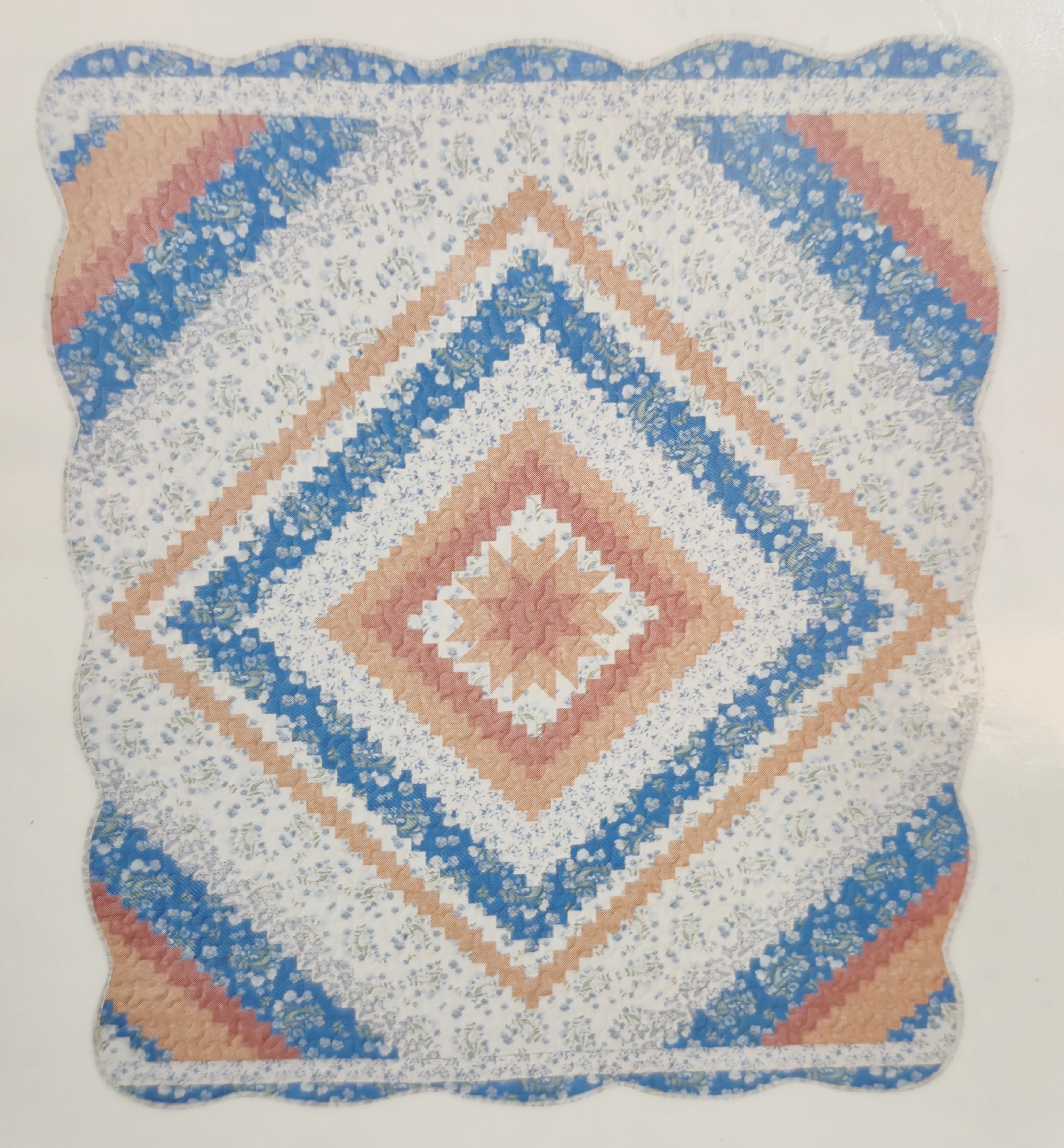 Peach, Blue and White Diamond Pattern Quilt  - A cheerful quilted pillow in a diamond patchwork pattern brings a warm, handmade touch to housewarming gifts and casual home decor. This decorative arrangement is perfect for someone who loves cozy, vintage-inspired accents-the person you're thinking of will notice the soft scalloped edge, the gentle palette of blue, peach and white, and the small floral print that livens the lighter bands.  The square cushion features stitched diamond quilting that adds texture and a craft-made feel; alternating bands of blue, peach and white create a symmetrical geometric pattern framed by a subtle scallop. The colors read as bright but slightly nostalgic, balanced by small floral motifs on the lighter strips and a neat finish that suits everyday sofas, entry benches, or bedroom nooks. Choose the Deluxe option and we will mount the quilt on a simple metal easel for an additional $15 perfect for display at a service or reception. Sized and styled for easy decorating, it's a comforting accent that makes recipients feel genuinely seen-an ideal pick for housewarmings, hostess gifts, or anyone who appreciates handmade-style home accents.