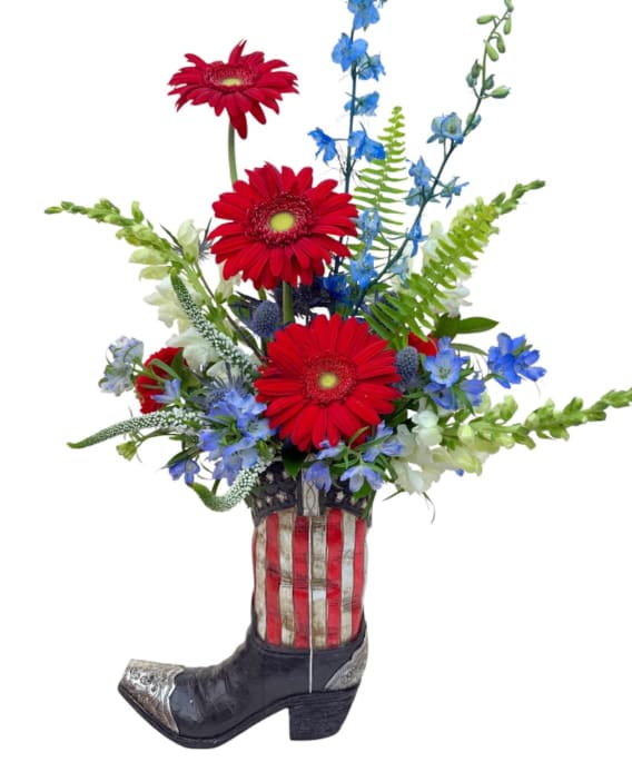Sophia's American Cowboy Boot - Red and white tulips burst from a novelty cowboy boot vase painted with an American flag motif, making this flower arrangement a playful, patriotic choice for Fourth of July, Memorial Day, or an all-American birthday bouquet. The bold contrast of bright tulip blooms against the dark boot gives a fresh, cheerful energy-perfect for the person who loves country charm with a spirited twist.  Tightly clustered red and white tulips form the heart of the design, accented by mixed-color touches that add depth and a casual, handcrafted feel. Displayed in a decorative cowboy boot container rather than a traditional glass vase, the piece measures as a compact, gift-ready arrangement that's easy to place on a mantle, entry table, or beside a celebration cake.  This is an ideal pick for anyone who enjoys rustic novelty and patriotic flair-send it to show appreciation, celebrate a milestone, or brighten someone's day with something unexpected. Care is simple: keep water fresh and place away from direct heat to extend the life of the tulips.