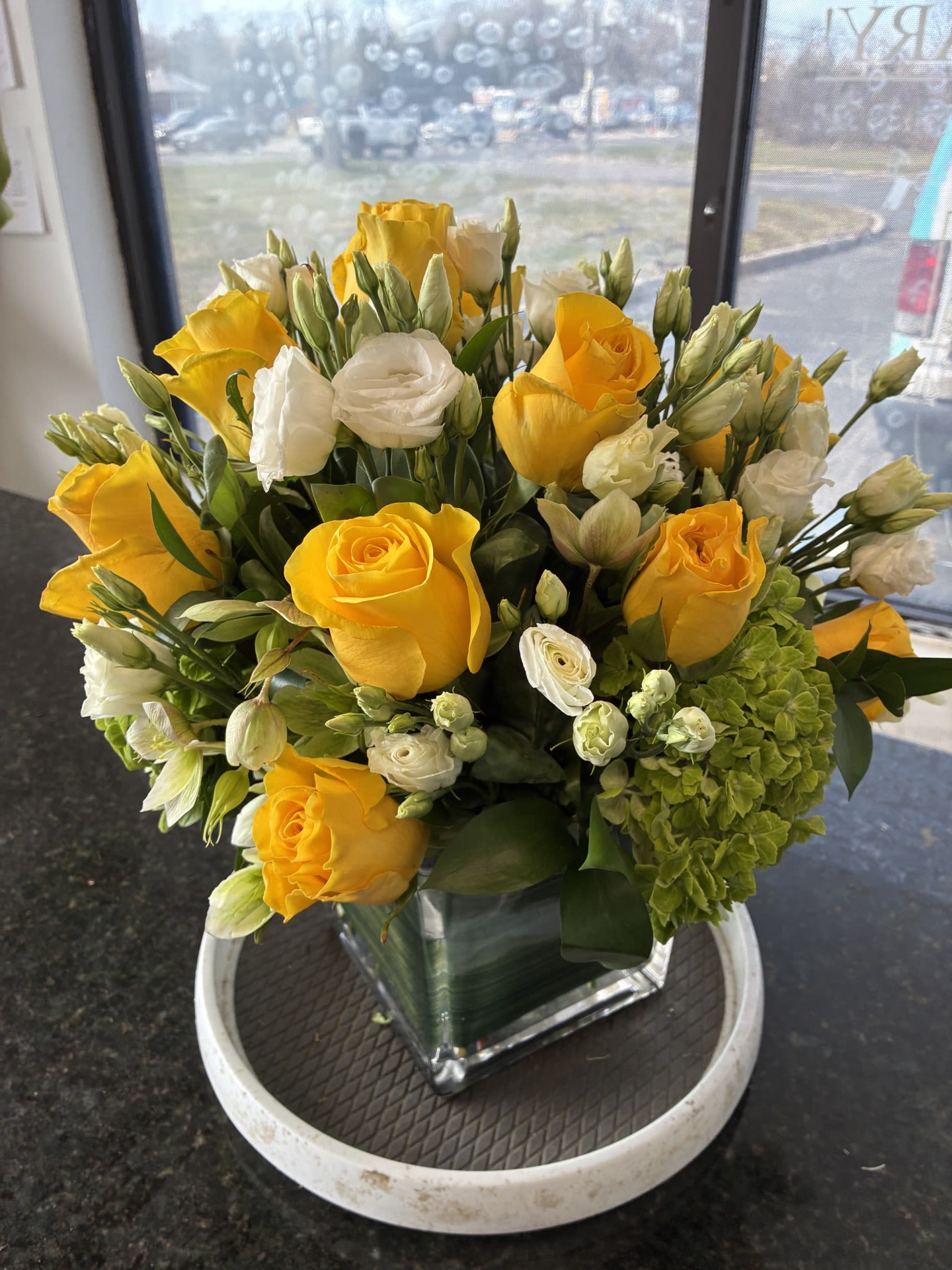 SUN-SUN-SUN - Yellow and Green..............For that Table centerpiece or just to say "Happy Easter"....standing about 16 inches tall and 14 inches Wide, made in a 51/5 in Glass lined Cube , with Yellow Roses , White Lisianthus and green Helleborus.
