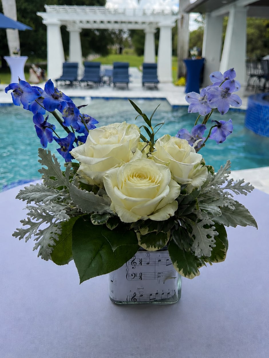 Melody Garden - A custom-designed floral arrangement featuring elegant white roses, fresh greenery, and soft blue delphinium accents, styled in a glass vase with musical note detailing for an artistic and personalized touch.  Best For: Graduation Celebrations, Recitals &amp; Performances, Birthday Gifts, Music-Themed Events, Teacher Appreciation, Dance &amp; Arts Celebrations, Personalized Gifts, Coastal &amp; Poolside Events, Sympathy &amp; Thinking of You, and Custom Event Decor.  This arrangement can also be customized with different flowers, colors, photos, or themes to match your occasion.  Flowers and accents may vary based on availability while preserving the overall style, palette, and quality of the arrangement.