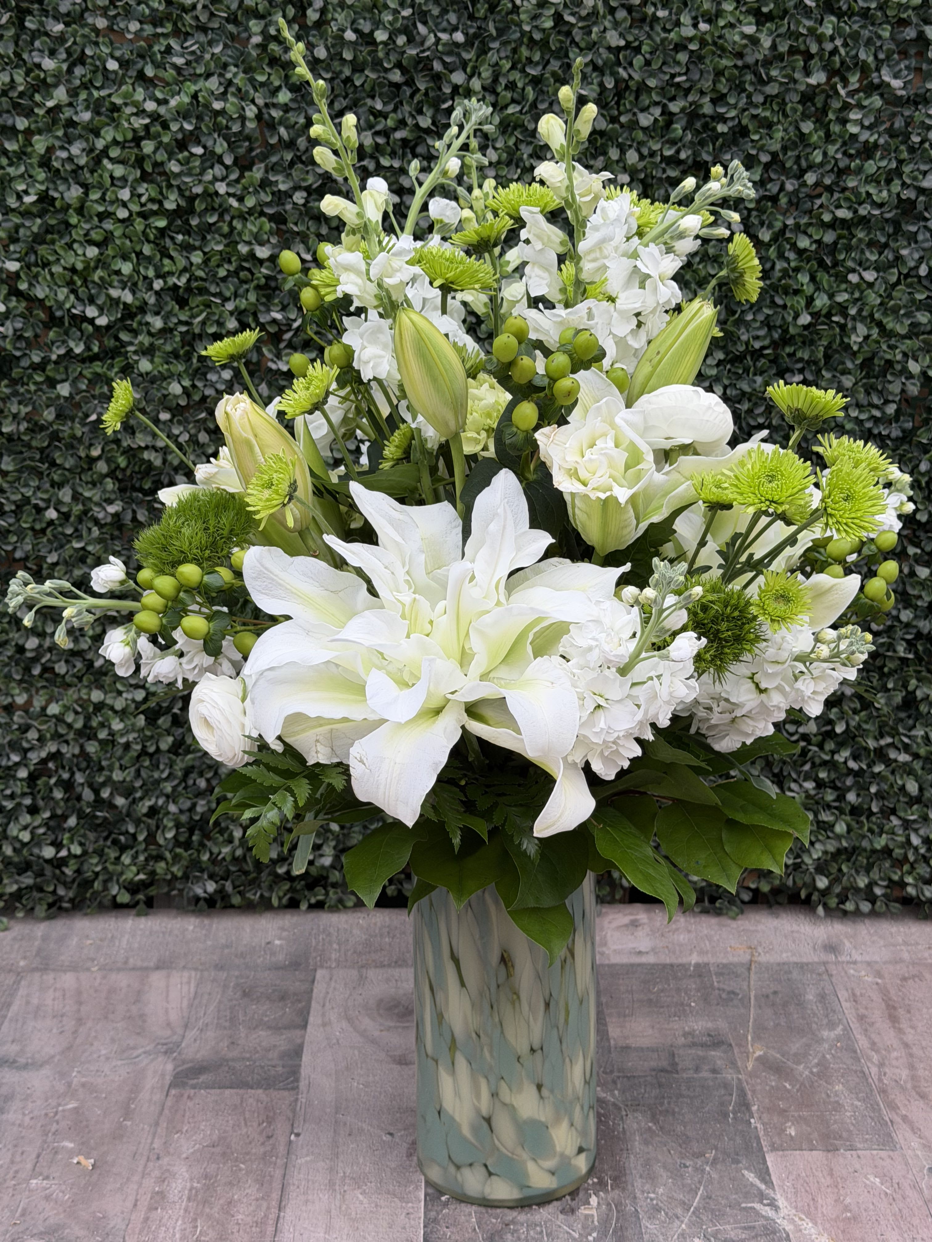Tall white and green floral arrangement with lilies and mums in a patterned glass vase
