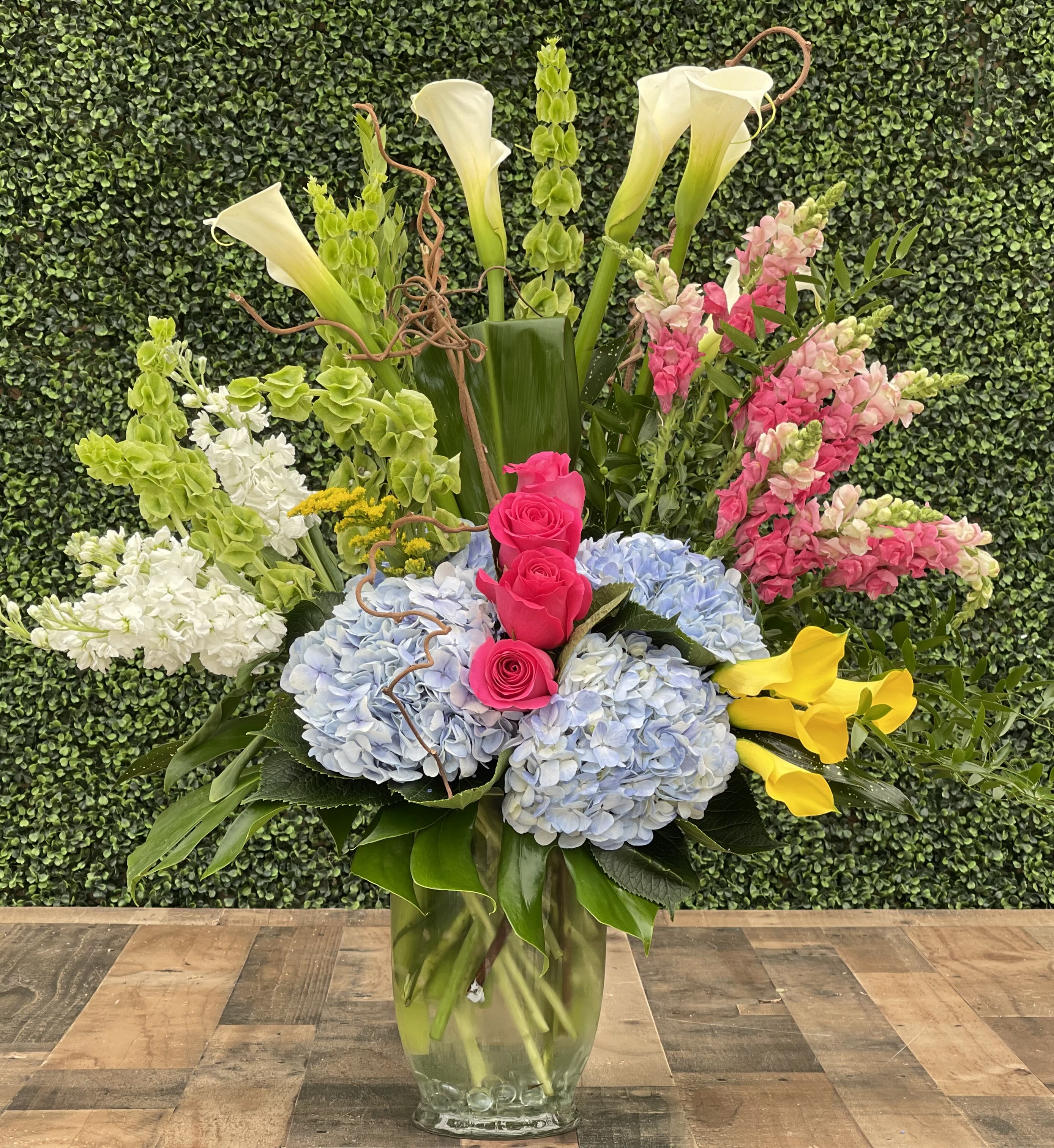 Tall mixed floral arrangement in a glass vase with hydrangeas, roses, calla lilies, and snapdragons