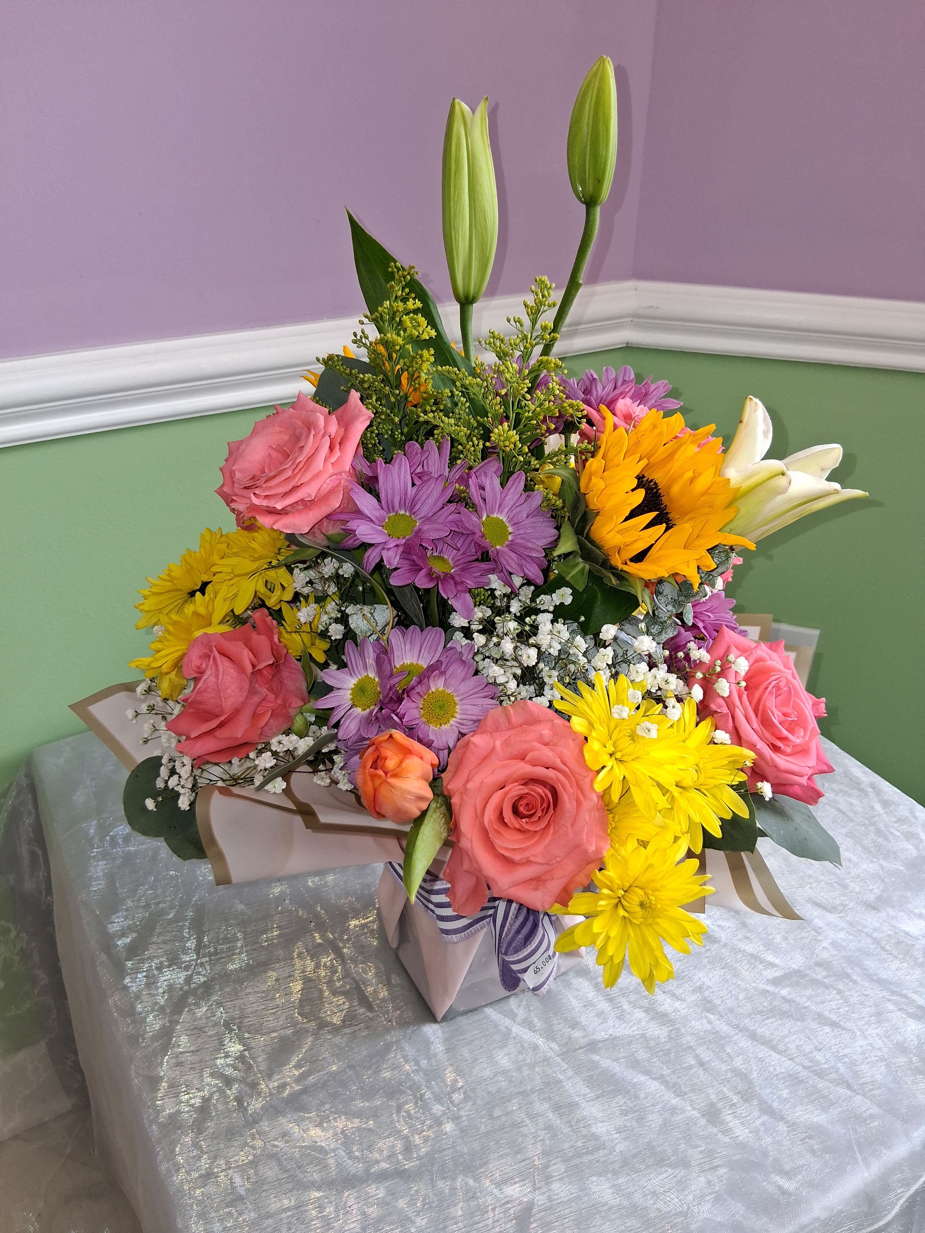Vibrant Table Arrangement  - A bright, cheerful centerpiece that reads like a celebration in bloom, this flower arrangement is perfect as a birthday bouquet, congratulations gift, or an upbeat thank-you. The compact gift-box presentation and lively mix of pink roses, yellow daisy-like blooms, and purple chrysanthemums makes the recipient feel truly seen-joyful, noticed, and celebrated the moment they open the box.  Dense, upward-reaching layers create a full, abundant silhouette: soft pink roses and unopened lily buds add romantic height, yellow daisies bring sunny cheer, purple mums introduce rich contrast, and white baby's breath lends a delicate, airy finish. The arrangement arrives wrapped with decorative paper, tied with a ribbon, and placed in a stylish gift box for easy presentation.  This is the kind of arrangement that suits friends, coworkers, or family when you want to brighten someone's day or mark a happy milestone. Order with confidence for same-week gifting; light daily water refreshes for any stems that may arrive cut, and keep out of direct heat to prolong bloom life.