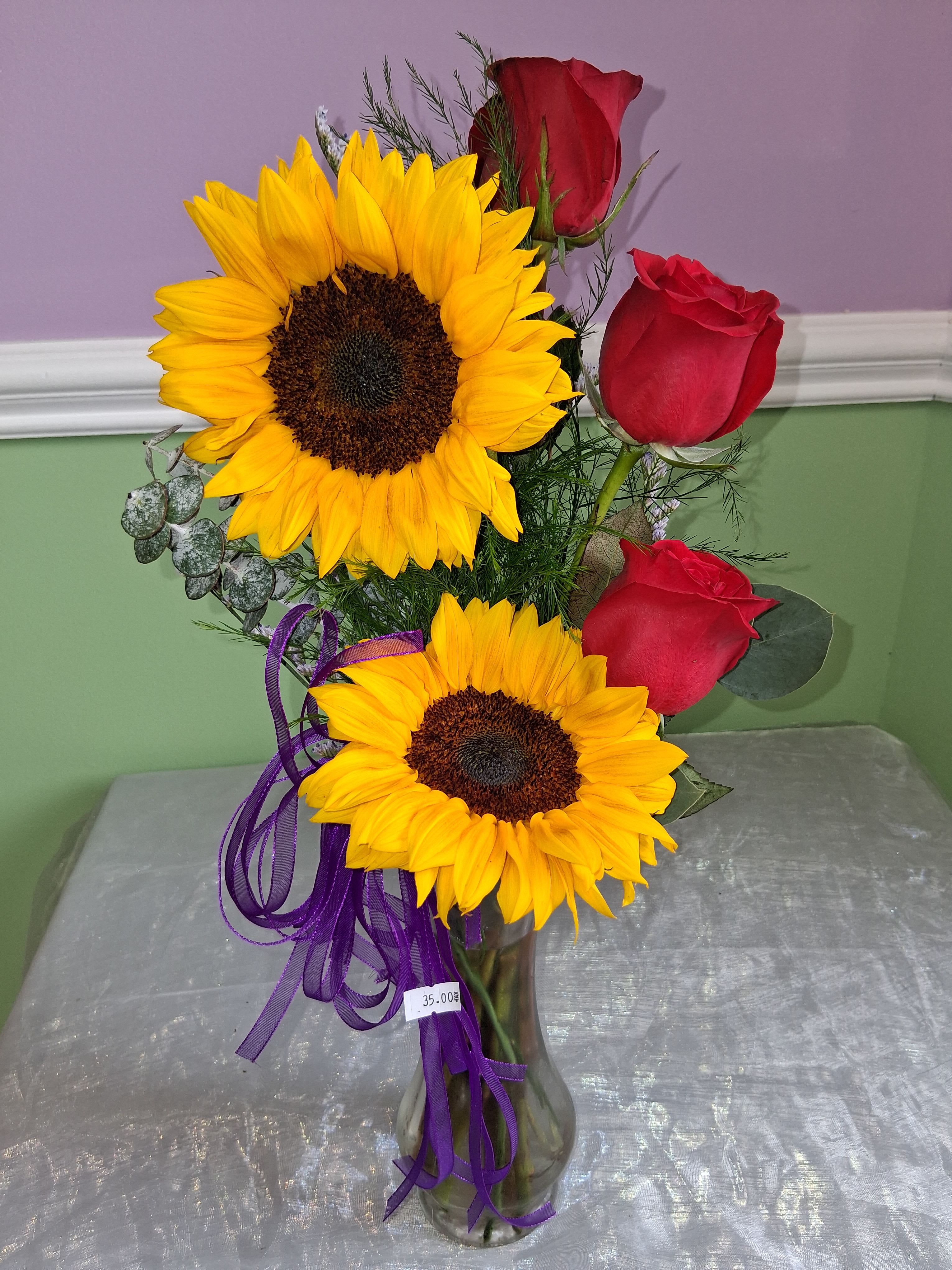 3 Roses with sunflower - Two large yellow sunflowers and three deep red roses create a bold, high-contrast birthday bouquet or romantic surprise that feels joyful the moment it's seen. This flower arrangement is ideal for birthdays, anniversaries, Valentine's Day, or "just because" moments when you want the person you're thinking of to feel both celebrated and cherished.  Arranged in a clear glass vase, the bright sunflower petals and velvety red rose blooms are complemented by fine filler greenery and a few silvery leaves for soft texture and movement. A ribbon tied at the base adds a pop of complementary color and a hand-tied, casual-gift charm; a discreet price tag is included on the vase. The scale is classic tabletop size-perfect for a desk, bedside table, or dining centerpiece-offering big visual impact without overwhelming a room.  Send this cheerful and romantic combination to make someone feel truly seen: it reads as both festive and intimate, a confident way to say "I'm thinking of you" or "happy birthday." For best enjoyment, place in fresh water and trim stems occasionally to extend vase life.