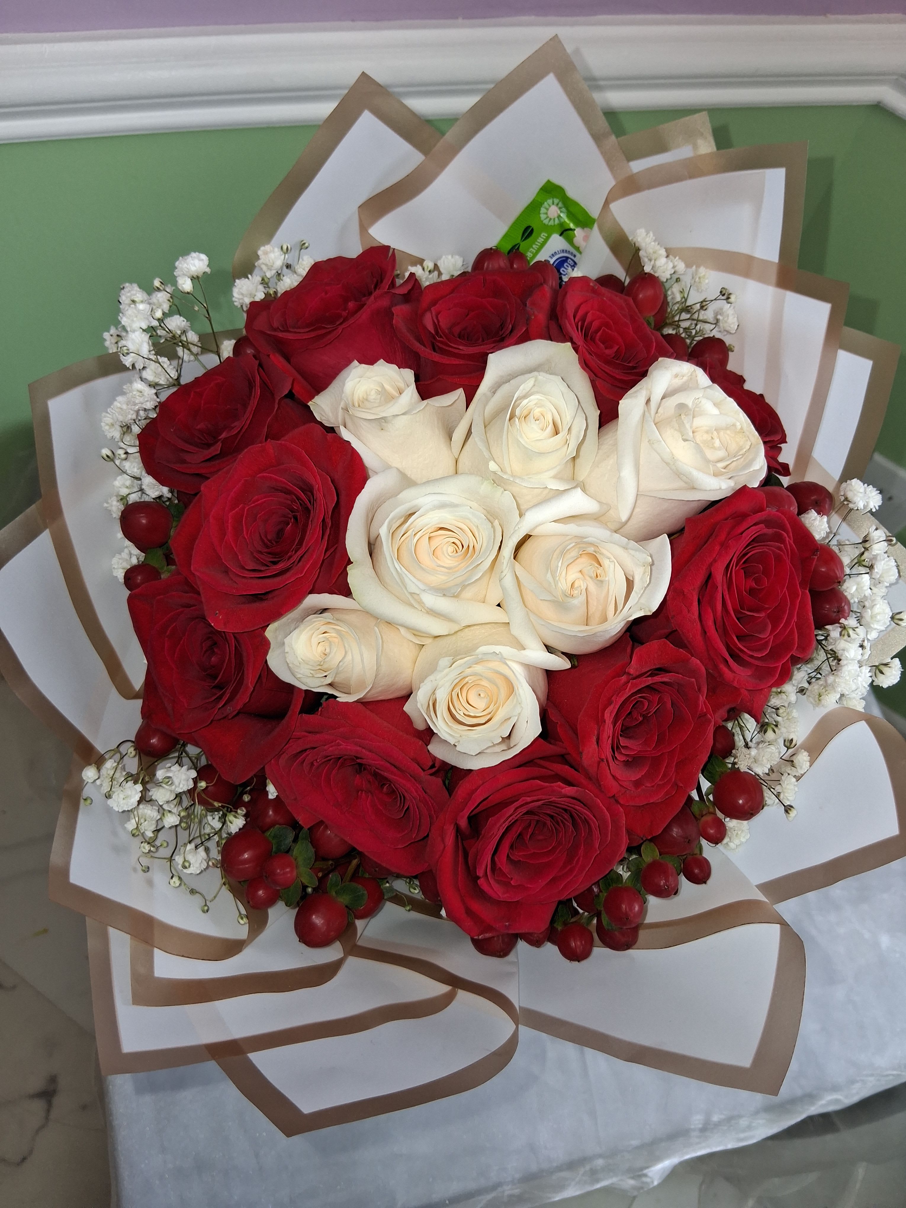 Red and White Roses 25 Roses - A striking ring of deep red roses encircles a creamy white heart at the center-this red and white roses bouquet delivers a classic, romantic statement perfect for an anniversary, Valentine's Day, or a wedding celebration. The high-contrast color story reads formal and timeless, so the person you're sending it to will feel cherished and truly seen.  Arranged in a dense, rounded cluster with delicate sprays of baby's breath softening the edges, the bouquet comes gift-ready wrapped in white paper with brown edging or gathered in a clear ribbed glass vase for a polished look. Finished with ribbon and decorative wrapping paper, it combines traditional roses with simple, elegant hard goods to create a memorable presentation.  Choose this arrangement when you want to say "I love you," "congratulations," or "thank you" with something refined and romantic; it's the kind of bouquet that makes anniversaries, proposals, or wedding moments feel elevated. Standard care: keep blooms in fresh water, trim stems every few days, and display out of direct sunlight to extend vase life.
