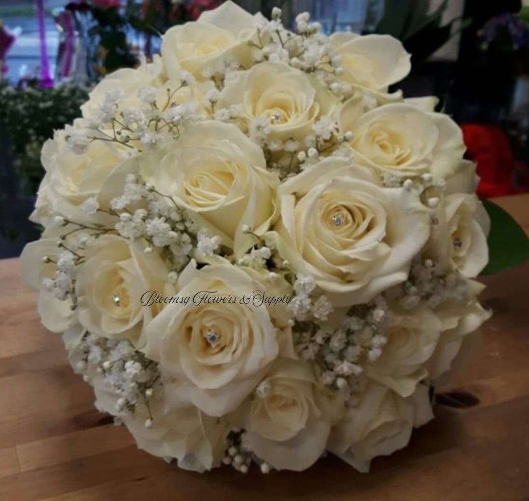 White Wedding Bouquet  - Cream-white roses gathered into a rounded, hand-held bouquet create an instantly romantic bridal bouquet or sympathy arrangement that reads as timeless and elegant. The pale monochrome palette and formal shape make this wedding bouquet a perfect choice for a classic ceremony or a respectful, understated sympathy gift for the person you're honoring.  Tightly packed rose heads are softened by delicate clusters of baby's breath tucked throughout, giving the bouquet an airy texture against the smooth, velvety petals. The compact, rounded silhouette is easy to carry and photographs beautifully; its neutral tones pair seamlessly with most color schemes and decor.  Whether you're marking a wedding, anniversary, or offering condolences, this bouquet conveys calm, thoughtfulness, and refined taste-she'll feel genuinely seen and comforted. Sized as a hand-held posy, it arrives ready to present; keep stems in water until the moment it's needed to preserve freshness.