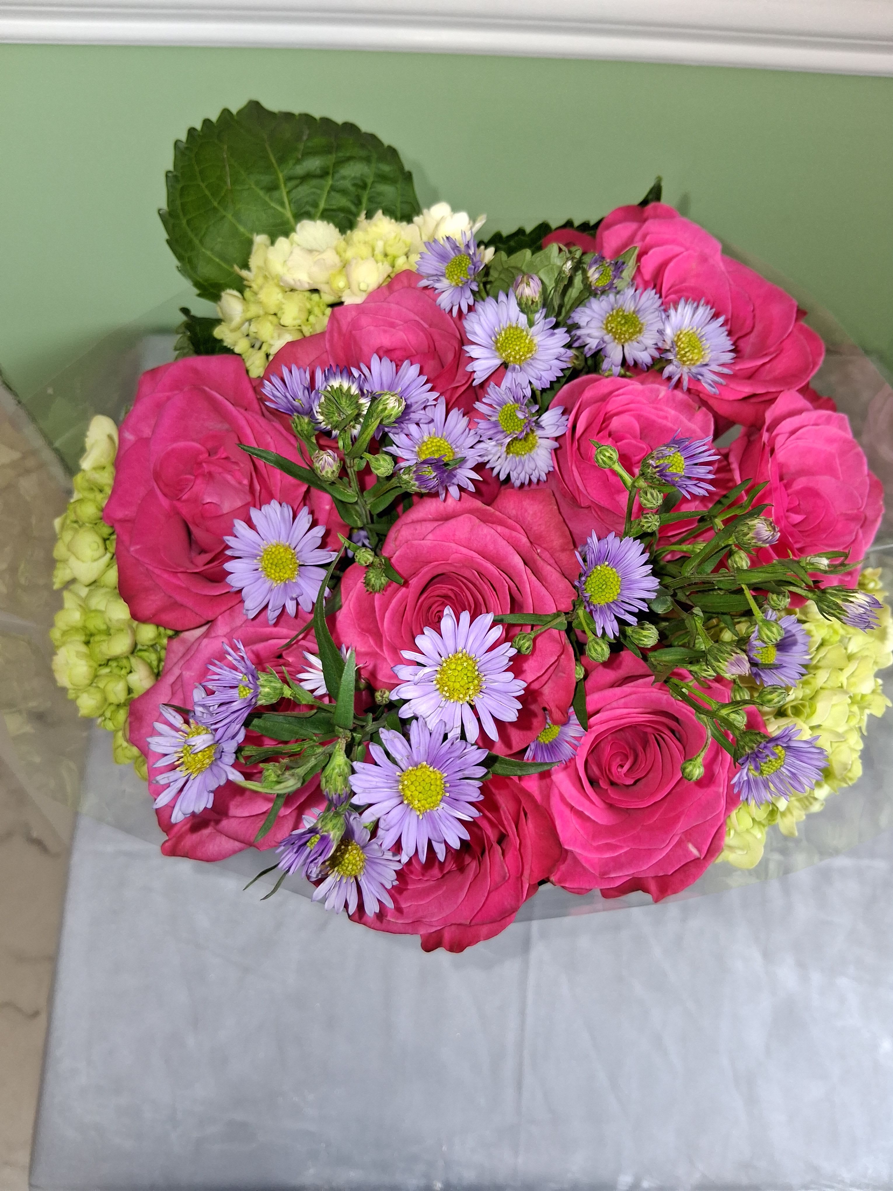 Pink Roses with Monte casino and Hydrangeas  - Vivid hot pink roses take center stage in this romantic flower arrangement, perfect as a birthday bouquet, anniversary flowers, or a love-and-romance gift. The bright pink blooms are contrasted with small lavender aster (Monte Casino) accents and pale green hydrangea clusters, creating a layered, full look that reads fresh and celebratory. The mix of saturated pinks and soft purples gives the recipient an immediate sense of joy and being cherished.  Tightly gathered into a rounded, hand-tied shape, the arrangement is framed by a large dark green leaf for depth and visual balance. It arrives wrapped in clear plastic and silver-toned paper for a polished presentation, and is available placed in a tall clear glass vase so it's gift-ready on arrival. This is the kind of bouquet that makes someone feel truly seen-bright, elegant, and thoughtfully assembled for special moments.