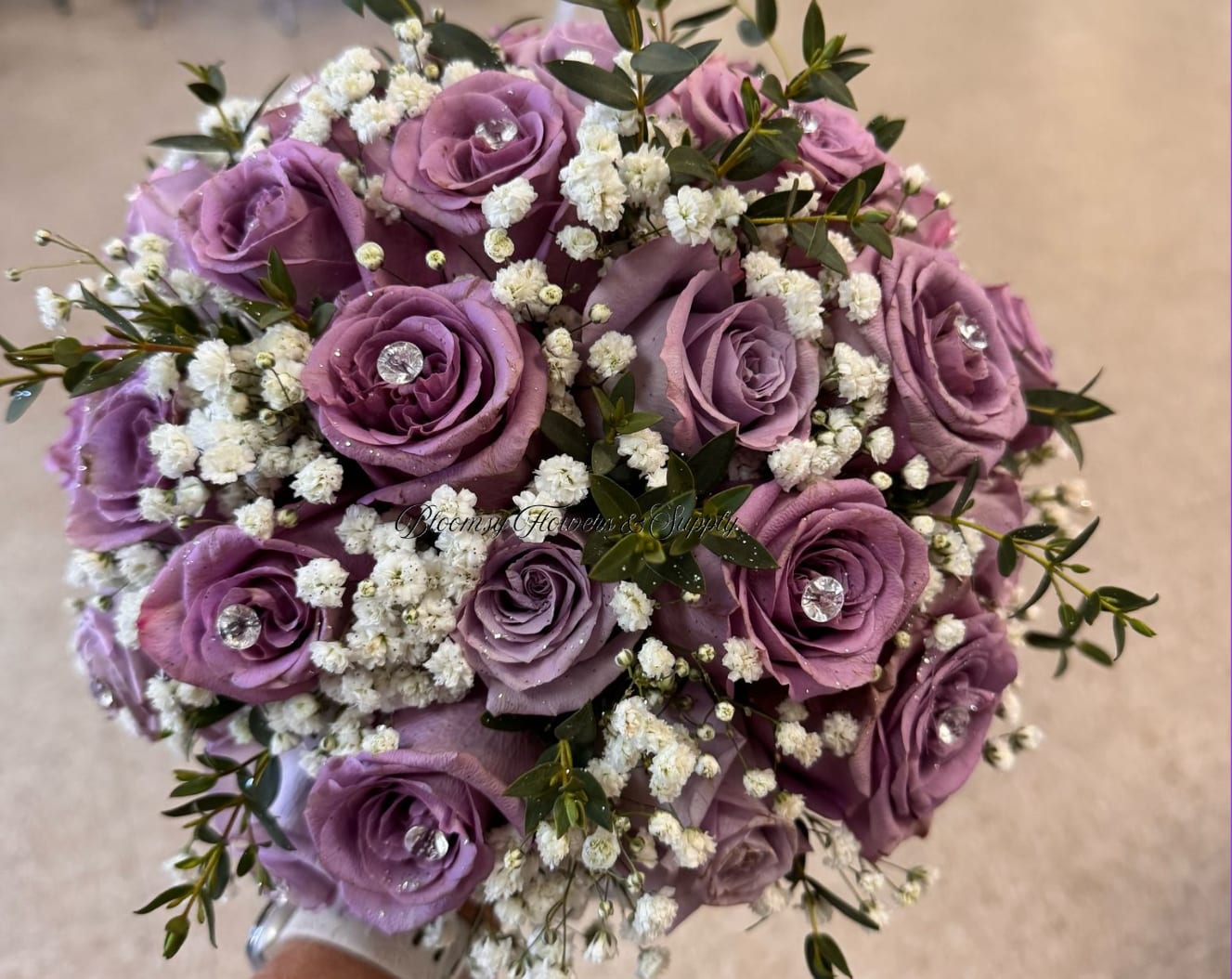 Wedding Bouquet  - Lavender roses clustered into a neat, round wedding bouquet create a soft, romantic statement perfect for a wedding bouquet or anniversary bouquet. The cool purple tones paired with touches of white and green set a gentle, dreamy mood that will make the person you're thinking of feel cherished and celebrated on their special day.  This hand-tied arrangement features tightly grouped lavender roses accented with delicate white baby's breath and sprigs of dark green foliage for contrast and texture. Small clear gem-like accents tucked into several rose centers add a subtle, sparkling detail that catches the light without overwhelming the design. The compact, round silhouette is easy to carry and photographs beautifully from every angle.  Ideal for brides, bridesmaids, or as a romantic gift to mark an anniversary, this bouquet reads classic and refined while feeling quietly modern. Order based on your wedding colors or gift intent, and expect a polished, ready-to-hold bouquet that will stay fresh with standard floral care: keep stems in water until the moment of presentation and store in a cool spot away from direct sun.