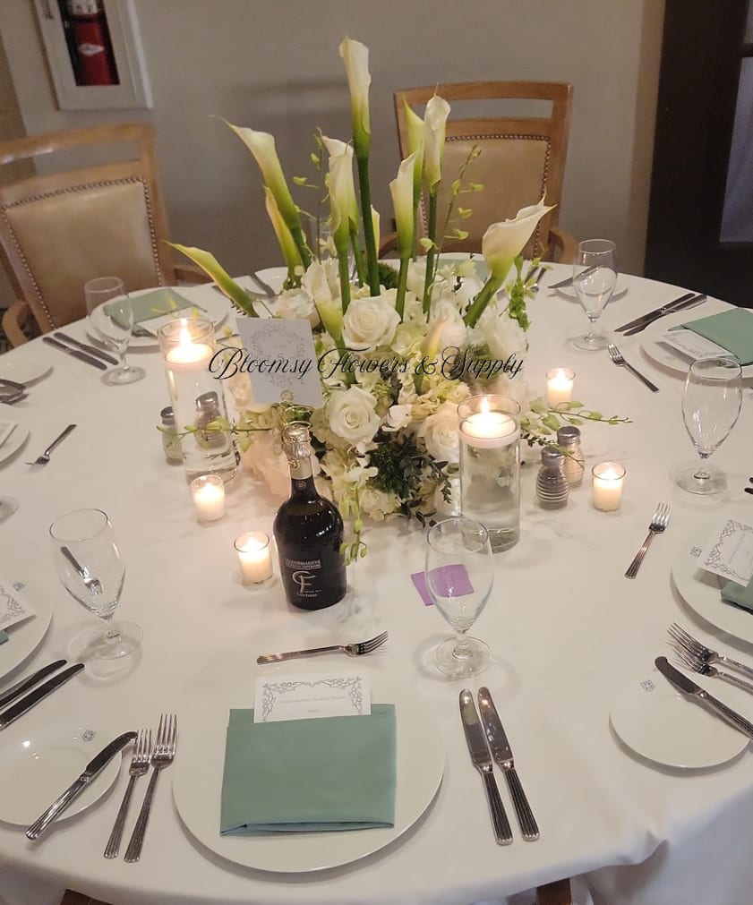 Calla Lillies Table Arrangement  - Tall white calla lilies soar above a lush base of white roses and petite white accents to create a formal centerpiece perfect for a wedding centerpiece or upscale dinner reception. The all-white palette with soft green stem accents reads as timeless and romantic-the kind of arrangement that makes the person you're celebrating feel honored and truly seen.  Presented in a clear cylindrical glass vase, the long, architectural calla stems and open blooms create a striking vertical line while the dense ring of roses adds softness and fullness at table level. Shown styled with glass votives, candles, wine glasses, place settings, and crisp linens, this arrangement is sized to crown a round dining table without blocking conversation-an elegant focal point for rehearsal dinners, anniversary celebrations, or any refined event where classic white florals set the tone. Care: keep water fresh and stems trimmed for lasting beauty.