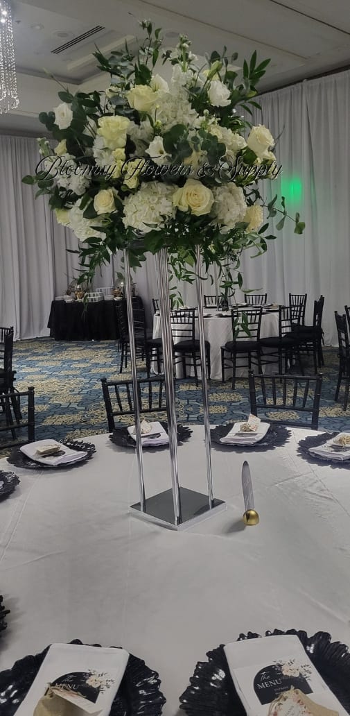 White center piece  - A towering white centerpiece designed for weddings or upscale receptions, this event arrangement crowns a chrome stand with a dense cluster of white roses, white hydrangeas, and delicate white accent blooms. The clean white-and-green palette reads formal and refined-perfect for a bride's table, head table, or any celebration where you want the person you're honoring to feel celebrated and seen.  Dark green foliage fans around the blooms to create a full, airy silhouette that reads beautifully from across the room and against neutral draped walls. Shown as part of a coordinated banquet setting with table linens, black charger plates, menu cards, napkins, and banquet chairs, the design complements classic linens and modern place settings without competing for attention.  Made to elevate event tables, this tall centerpiece brings height and elegance to wedding receptions, anniversary dinners, or milestone parties. Because it sits on a chrome stand, the floral mass stays visible while leaving tabletop space for place settings; standard care-fresh water and occasional misting-keeps roses and hydrangeas looking their best through the day and evening.