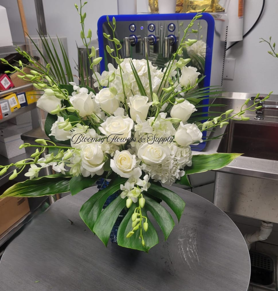 Elegant Table Wedding Arrangement - A refined cluster of white roses and white hydrangeas framed by arching green Phalaenopsis orchids creates an elegant wedding bouquet or sympathy arrangement that feels both serene and sophisticated. The clean white-and-green palette speaks to moments that call for quiet beauty-she'll feel comforted, honored, or cherished by the calming presence this design brings.  Dense, rounded heads of roses and hydrangeas form the visual core, while the glossy, broad leaves at the base give the arrangement structure and a layered silhouette. Delicate green orchid sprays arch above the cushion of blooms, adding graceful movement and a subtle textural contrast. The composition is presented in a bold blue vase that provides modern color contrast against the white flowers, making the arrangement suitable for formal events, memorial settings, or as a tasteful wedding centerpiece.  Perfect for expressing sympathy, celebrating a wedding, or offering a respectful tribute, this arrangement arrives ready to display in its blue vase. The sturdy vase and the compact, layered design make it easy to position on a reception table or beside a remembrance space; minimal care-fresh water and away from direct heat-will keep the blooms looking their best.