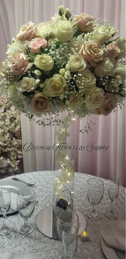 Rose Center Piece two tones pink and white - A tall, luminous centerpiece built from cream, ivory, and blush roses makes a romantic statement for weddings and elegant receptions. This rose arrangement for a centerpiece or event table reads soft and refined-the pale pink and white blooms form an abundant rounded top that will make the person you're celebrating feel truly honored and seen.  The design pairs tightly packed roses with airy white filler flowers in a clear glass vase wrapped with delicate string lights, adding a warm glow that lifts the arrangement into evening events. Shown styled with a tablecloth, candles, wine glasses and plates, it brings formal presence without feeling fussy; the vase height keeps sightlines open while the full floral dome reads lush from every angle.  Perfect for wedding head tables, anniversary dinners, or any reception where you want guests to pause and admire, this centerpiece arrives ready to place and shine. Care is simple: keep water fresh and out of direct, harsh sun; the string lights are low-heat for safe nightly ambiance.