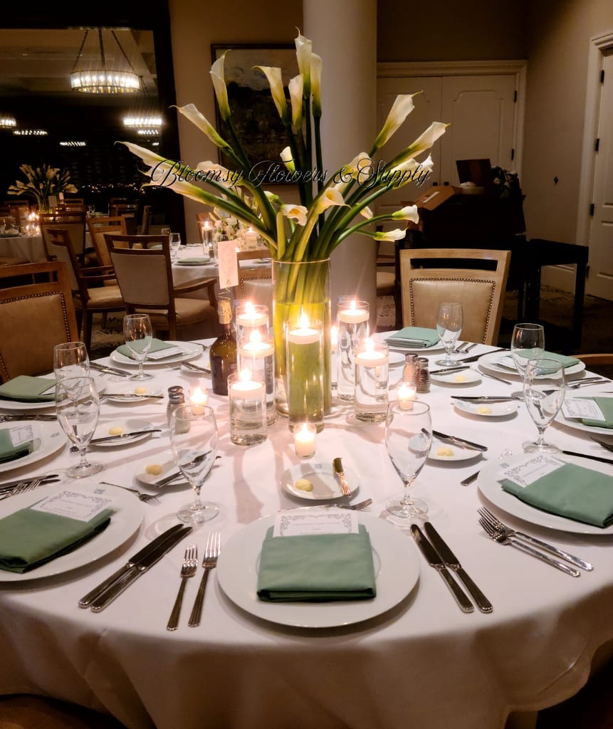 Tall Calla Lillie Arrangement  - Tall white calla lilies rise like sculpted candles from a clear cylindrical vase, making this floral arrangement an elegant centerpiece for a wedding reception or special dinner event. The arrangement reads as a formal, architectural statement-perfect for an anniversary dinner, rehearsal dinner, or any celebration where a refined, modern classic look will make the person you're honoring feel truly celebrated.  Long, upward-facing blooms and clean green stems give the bouquet a serene, sculptural silhouette that pairs beautifully with white plates, glassware, and folded napkins on a round banquet table. The clear glass vase keeps the focus on the lilies' smooth white spathes and glossy foliage; surrounding candles add a warm, intimate glow that softens the look and enhances the celebratory atmosphere.  Designed to sit at the center of a fully dressed table, this arrangement is sized to complement place settings without blocking sightlines, making conversations easy while still delivering visual impact. Minimal care is needed-keep water fresh and stems trimmed for lasting beauty through the event-so the person you're honoring can enjoy the moment rather than fuss over flowers.