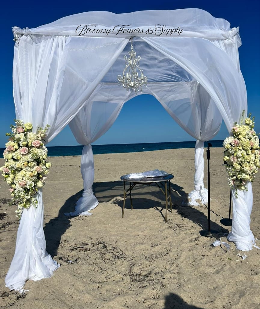 Tied Wedding Arrangement for multiple uses in ceremony  - This tied wedding arrangement pairs pale pink roses and creamy white blooms with touches of yellow and soft blue, designed to make the person you're honoring feel celebrated and seen as they stand beneath the canopy.  Large floral anchors at the front posts mix hydrangeas, roses, and complementary seasonal flowers for a full, textured look-lush clusters of petals against airy fabric and the shimmer of a hanging chandelier. The design works with metal stands and a table when needed, giving a polished, formal silhouette that still reads breezy and effortless for beach or outdoor weddings.  Ideal for couples planning a ceremony that blends elegance with seaside ease, this arrangement frames vows, photos, and first kisses with garden-fresh scent and soft color. Sizes are scaled for post anchoring and canopy use; please confirm site dimensions and installation needs when ordering so the florals and draping arrive ready to anchor your moment.