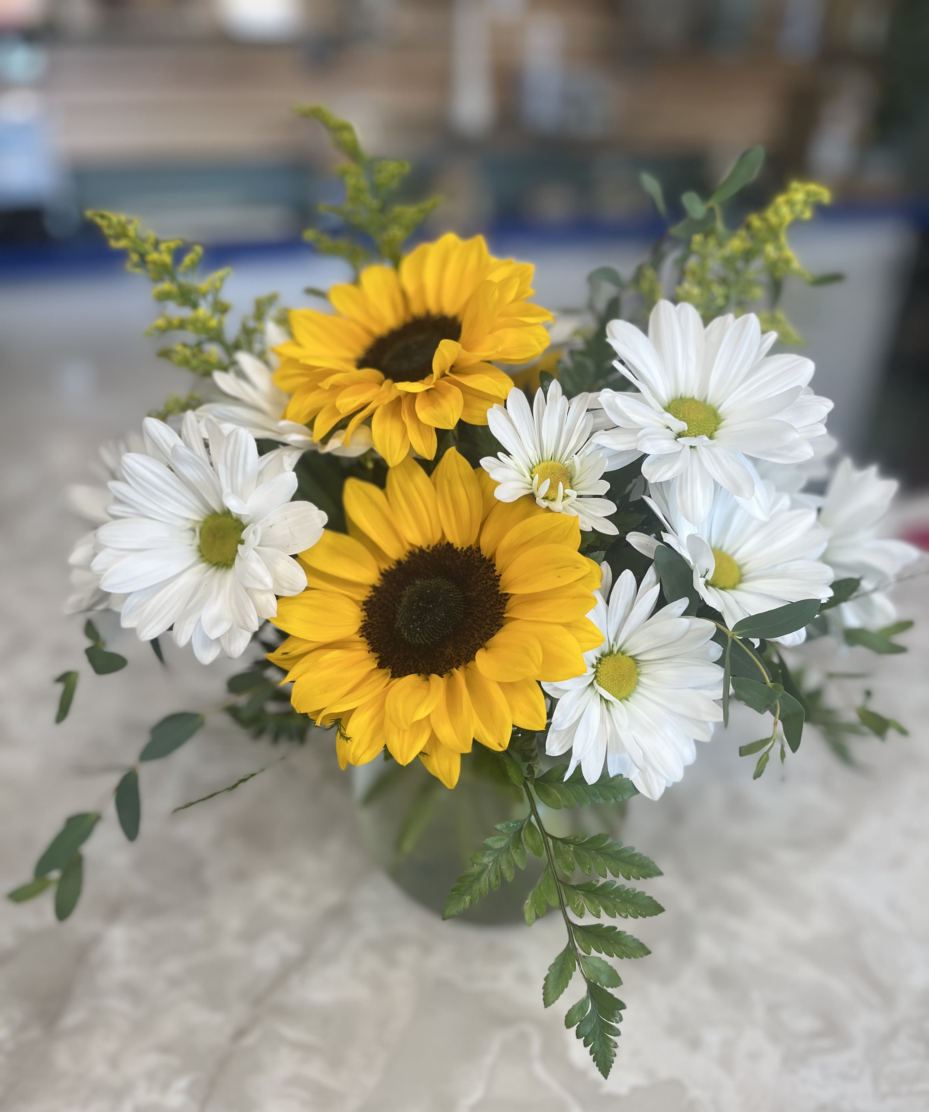 Sunny Days - This simple yet elegant all-around arrangement is the perfect centerpiece for any table this summer, with bright white daisies, glowing sunflowers, solidago and flowing greens arranged in a rounded vase!