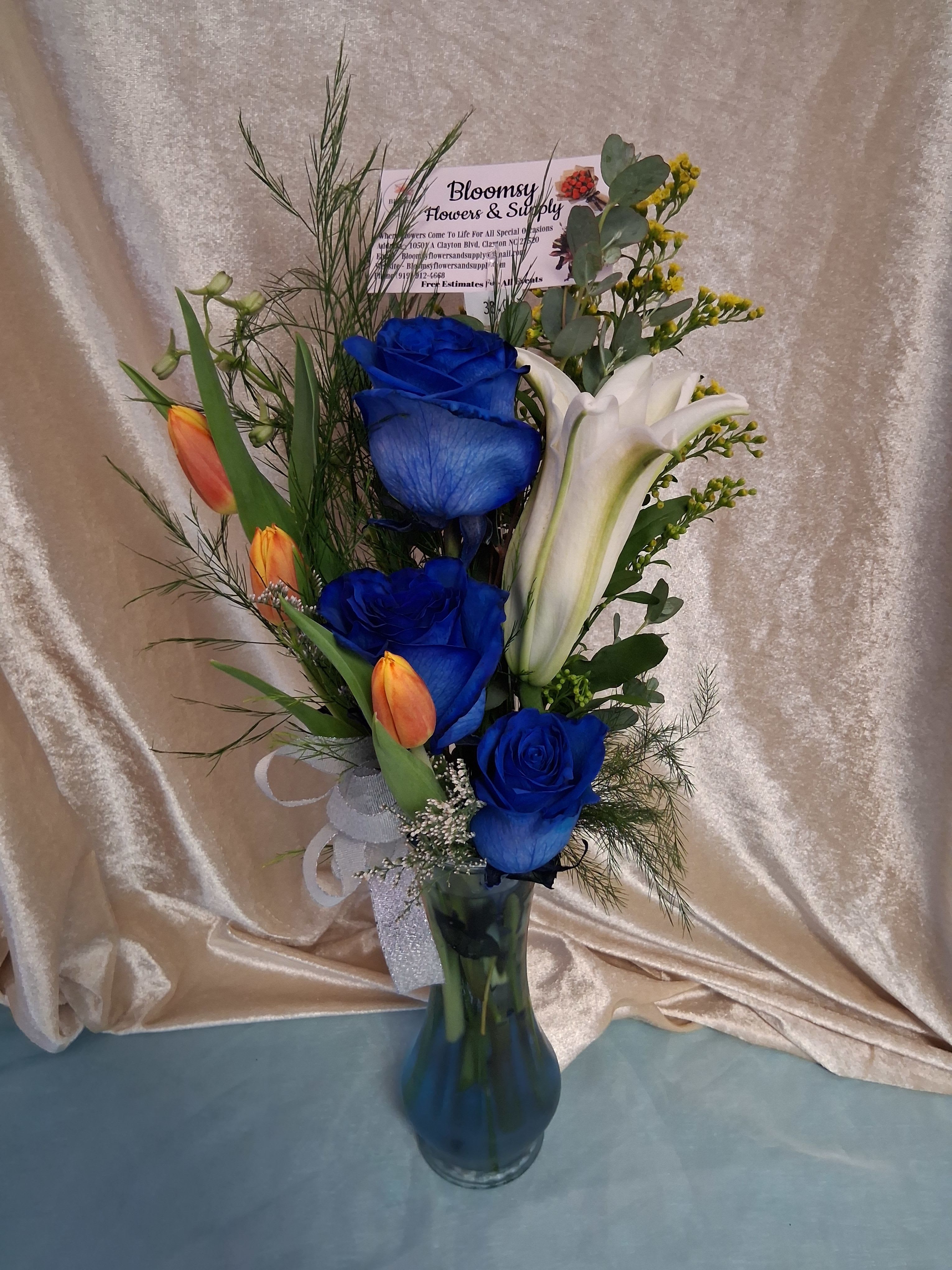 3 blue roses with tulips  - Vivid blue roses set the tone in this striking flower arrangement, their cool, saturated color balanced by a large white lily and bright orange tulips-a bold contrast that feels both elegant and slightly dramatic. Perfect as a birthday surprise, a romantic gesture, or a thoughtful "thinking of you" gift, this bouquet also suits Thank You, Father's Day, or Just Because moments when you want the recipient to feel genuinely seen.  The design includes three blue roses, a prominent white lily, and several orange tulips gathered with fine filler and airy greenery for height and texture in a loose, upright silhouette. The stems arrive arranged in a clear glass vase, finished with a pale decorative ribbon and a floral pick card tucked at the back for a personal message. Its mixed colors and refined presentation make it appropriate year-round for celebrations, appreciation, or a heartfelt hello.