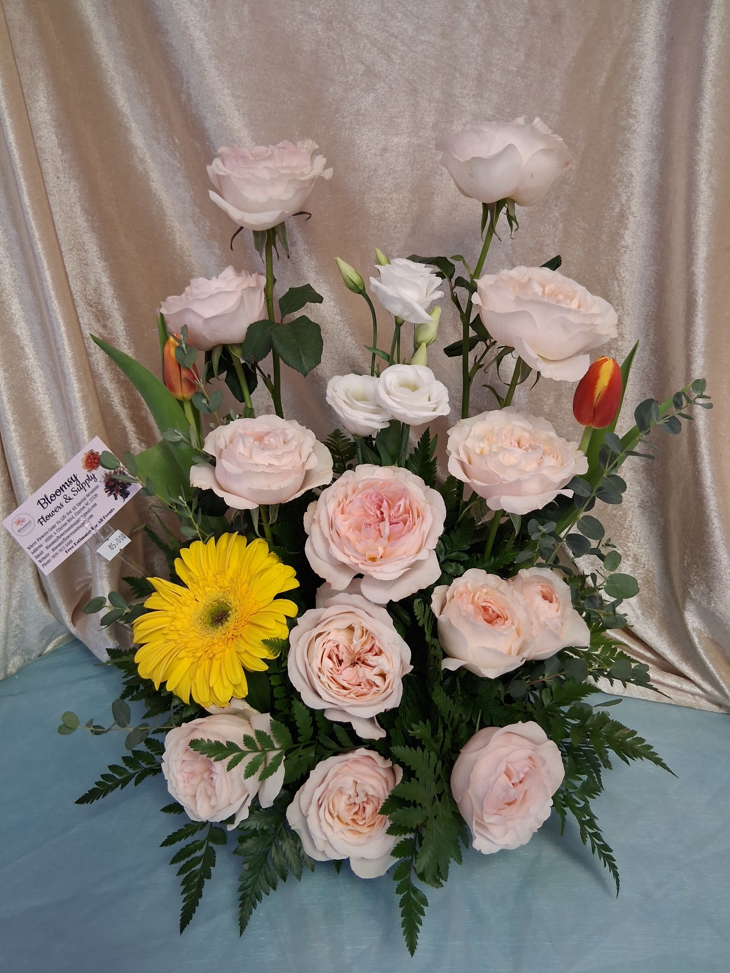 Pink Garden Rose Arrangement one dozen with mix flowers - Pale blush garden roses take center stage in this pink garden rose arrangement, a romantic bouquet perfect for anniversaries, birthdays, or a joyful springtime surprise. The tiered design places soft white tulips above a rounded cluster of one dozen roses, while a single bright yellow gerbera daisy pops near the lower left to catch the eye-an uplifting contrast that makes the recipient feel truly noticed.  Alongside the roses and tulips, airy stems and eucalyptus-like greenery give the arrangement a fresh, garden-style texture that reads both elegant and relaxed. Creamy pinks, soft whites, and touches of green create a gently romantic palette, with the tall, layered silhouette bringing graceful height and presence to any room. Presented as a floral arrangement with a price tag, this bouquet arrives ready to display and to make someone feel celebrated and cherished.