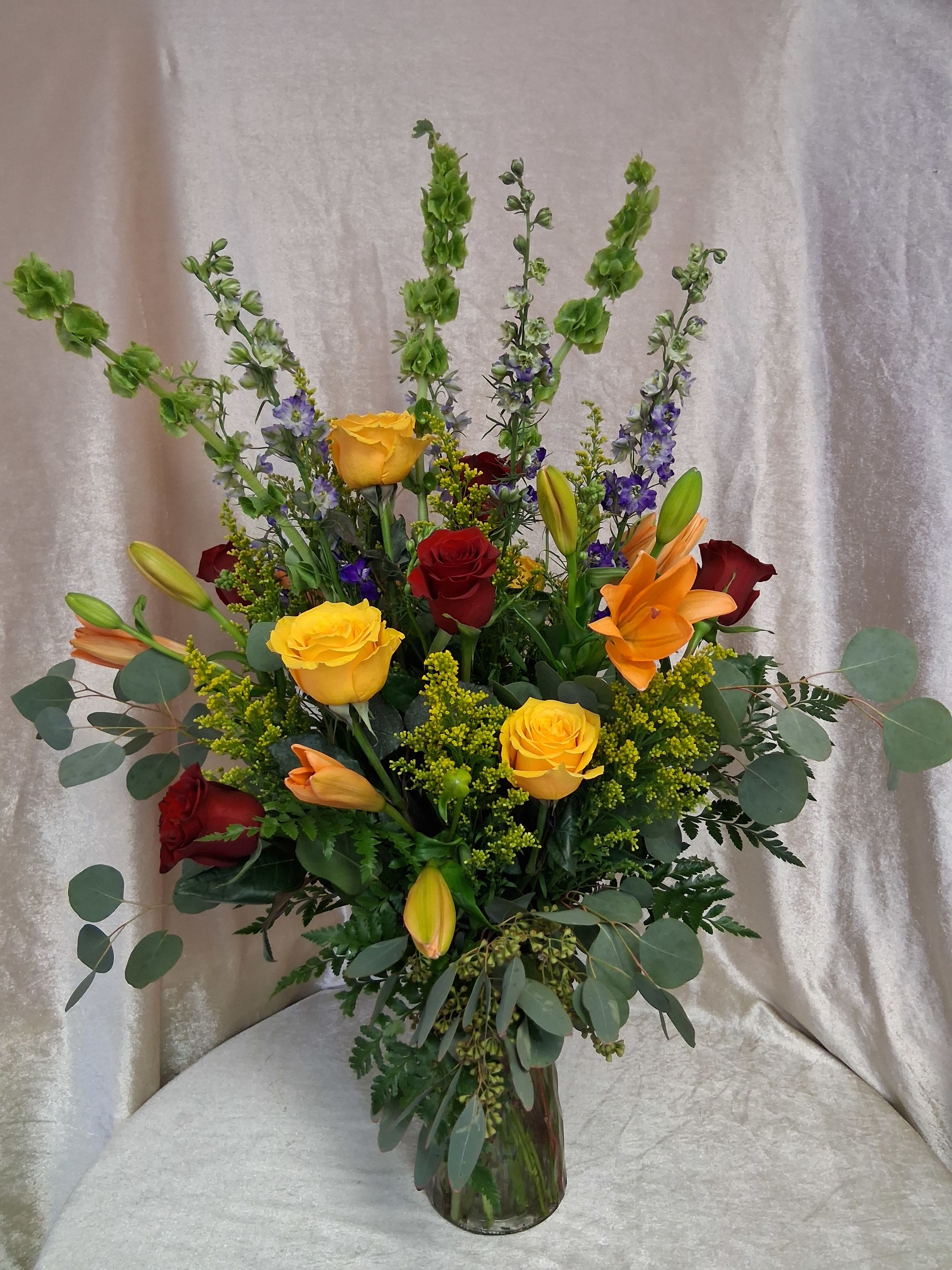 One Dozen Roses red and yellow with mix flowers - A lively twelve-rose arrangement that feels like a celebration in bloom - perfect as a birthday bouquet, congratulations gift, or a radiant romantic surprise. The mix of red and yellow roses reads both classic and joyful, while orange lilies and tall purple delphinium spikes add dramatic height and contrast to catch the eye the moment it enters the room.  Bright warm blooms are set among airy eucalyptus and assorted greenery for a layered, upright composition that's full without being heavy. The clear glass vase showcases the stems and keeps the look elegant and modern; colors include green foliage, sunny yellow, rich red, orange, and cool purple accents from the delphinium.  This is the kind of arrangement that makes the recipient feel truly celebrated - bold, festive, and thoughtfully composed. Arrives ready to display in the included vase; to extend bloom life, keep water fresh and trim stems every few days.