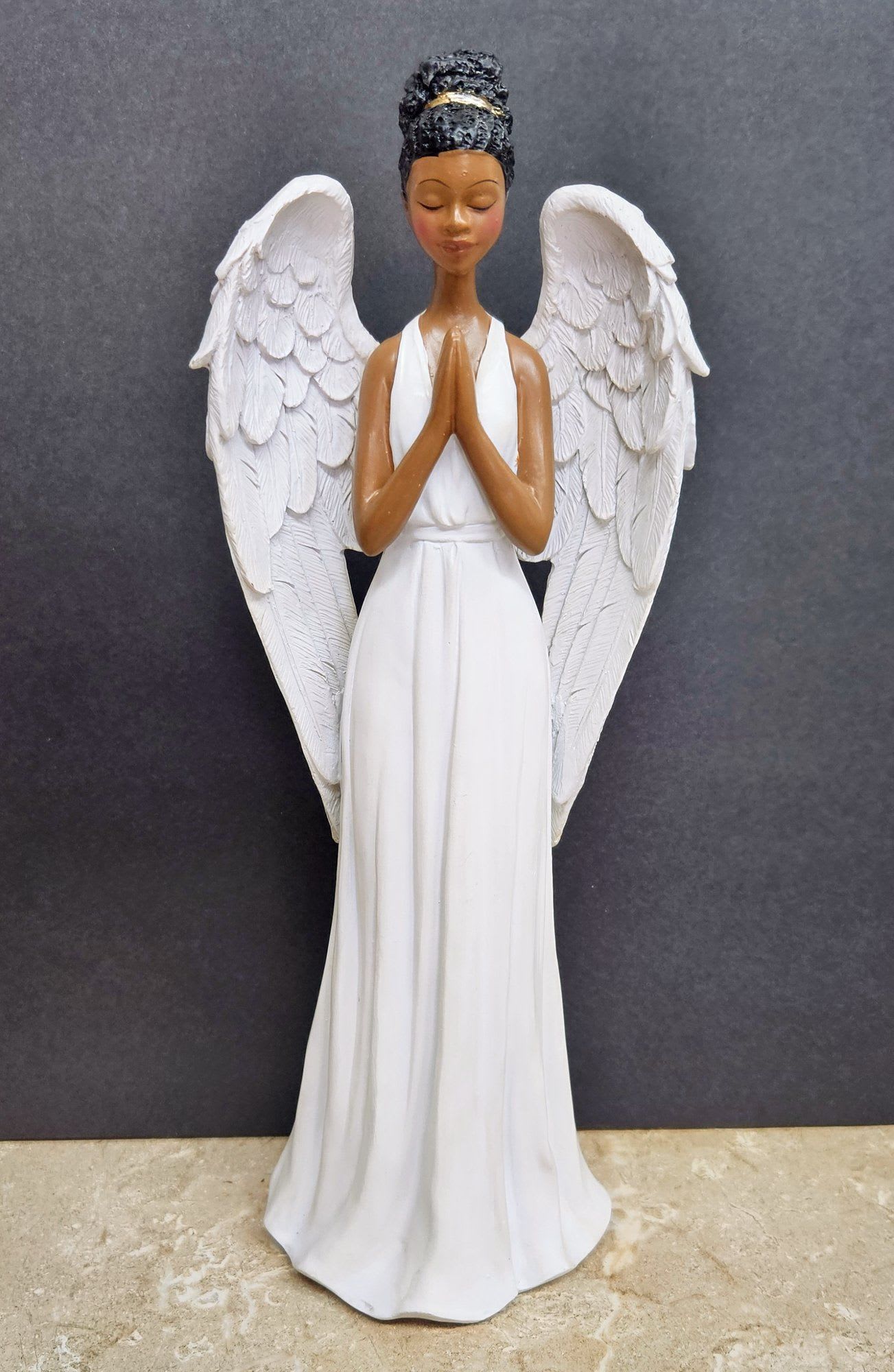 Angel of Grace - A serene standing angel figurine dressed in a long white gown, folded hands and large feathered wings - the perfect sympathy or funeral accent to let someone know you're thinking of them. The calm, prayerful expression and dark textured hair create a gentle contrast that reads devotional and comforting, offering a quiet focal point for remembrance or a peaceful corner of the home.  Handcrafted-feel sculpted details in the wings and dress lend texture and ornamentation without being ornate, so the piece works well in memorial settings, on a mantel, or beside a bedside lamp for daily reflection. Its neutral palette of white with dark accents pairs easily with fresh flowers or a simple votive; order with same-day delivery where available to ensure timely, thoughtful support for someone grieving or in need of comfort.