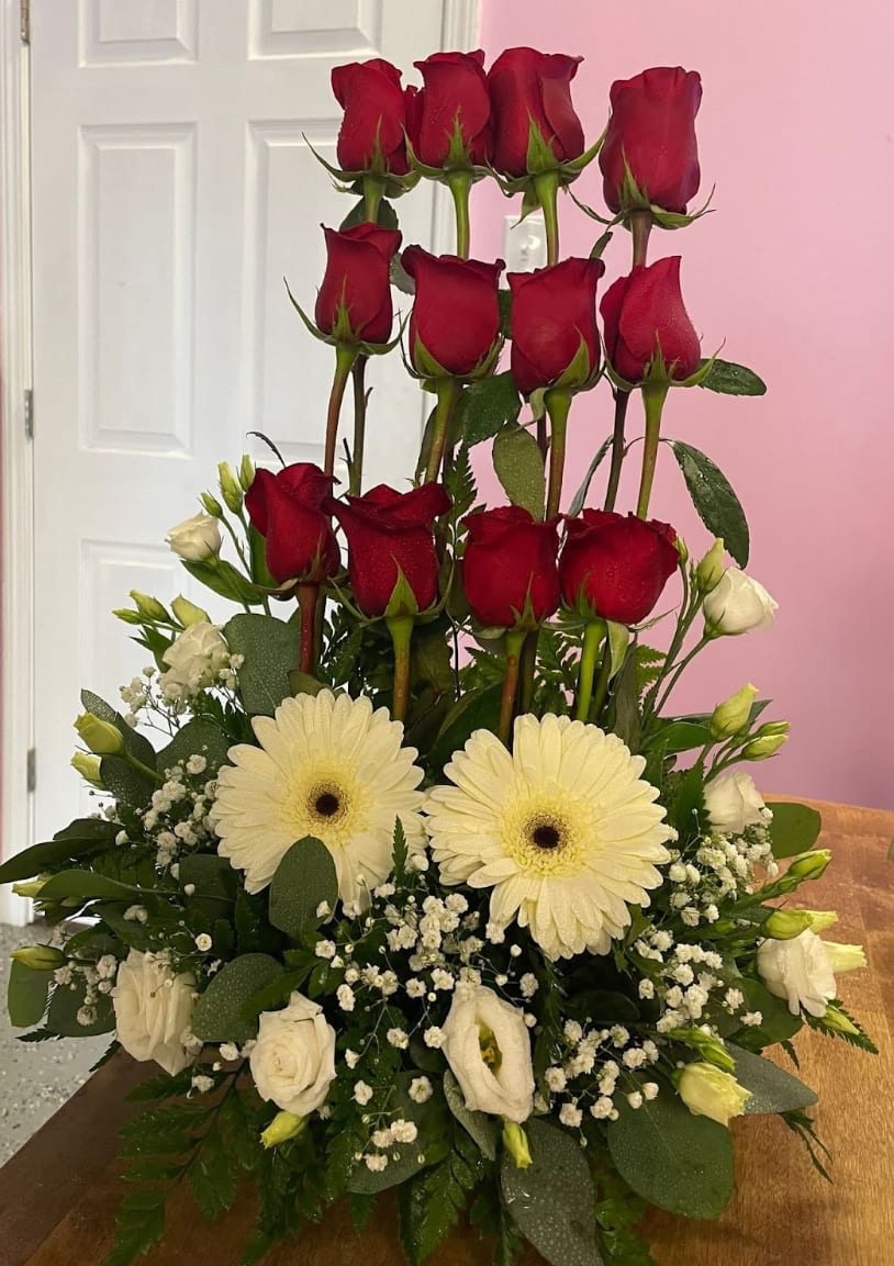 One Dozen red rose arrangement with white mix flowers - Deep red roses rise above a cloud of white blooms in this one dozen red rose arrangement-a romantic anniversary bouquet or a thoughtful sympathy arrangement that speaks with classic, heartfelt contrast. The vertical, layered silhouette makes a striking first impression: the red rose heads draw the eye upward while the white flowers below soften the composition, creating a dignified yet tender statement for the person you're honoring.  This tall design pairs a dozen long-stem red roses with white gerbera daisies and delicate white lisianthus (plus smaller white accent blooms) nestled into rich green foliage. Dense greenery fills and supports the lower half, giving the arrangement a full, substantial feel and clear structure. The color palette of red, white, and green emphasizes romance and reverence-perfect for anniversaries, expressions of love, or sending comfort.  Choose this bouquet when you want the recipient to feel truly seen-romanced, celebrated, or gently comforted-by a bouquet that balances bold emotion with soft elegance. The layered height makes it ideal for display in a tall vase or on a mantel; keep flowers in fresh water, trim stems every few days, and they'll maintain their visual impact for as long as possible.