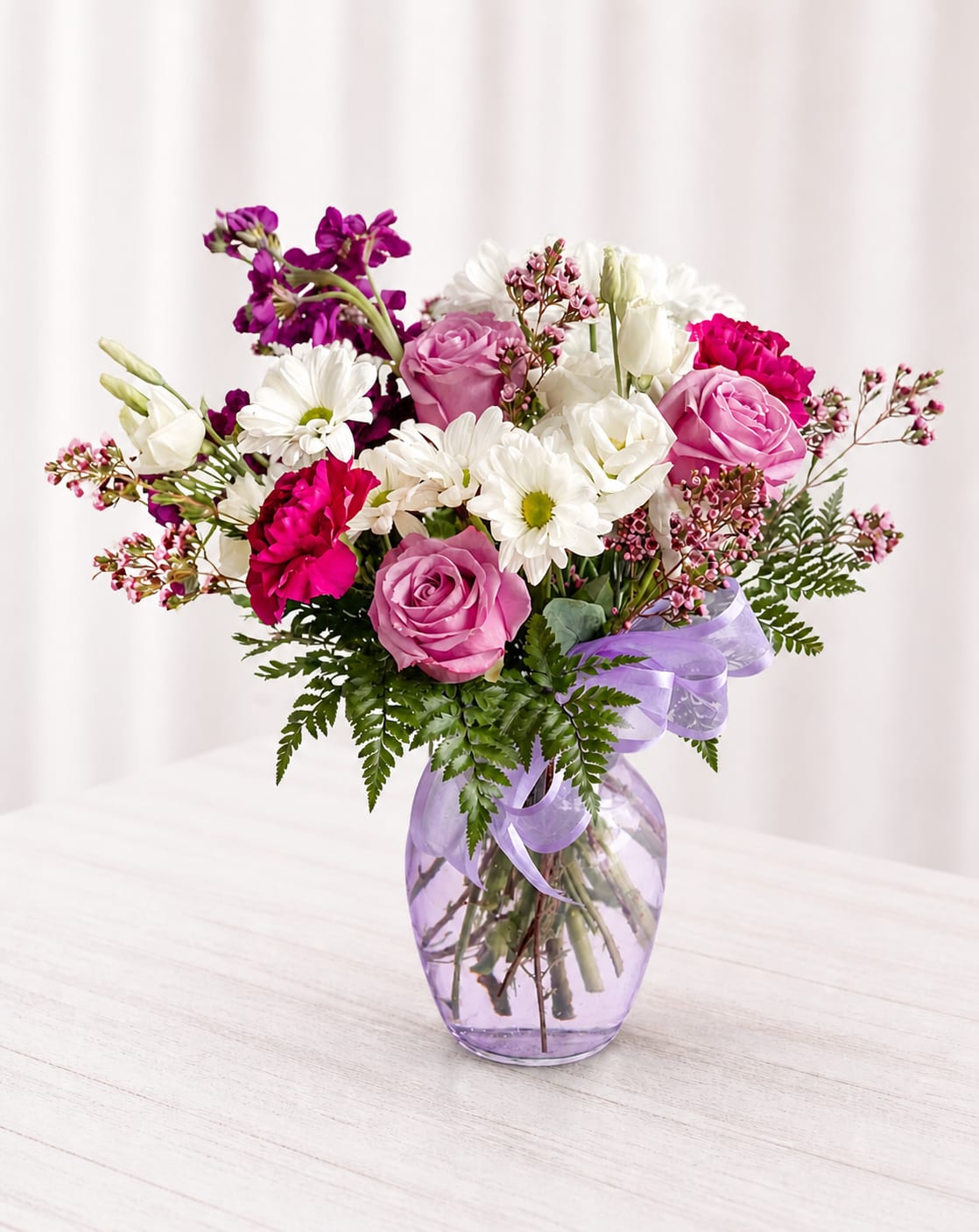 Virginia  - The Virginia is a mix of seasonal flowers in white and lavender. The mix includes roses, stock, carnations, daisies, and lisianthus in a lavender vase. Vase style may vary.  