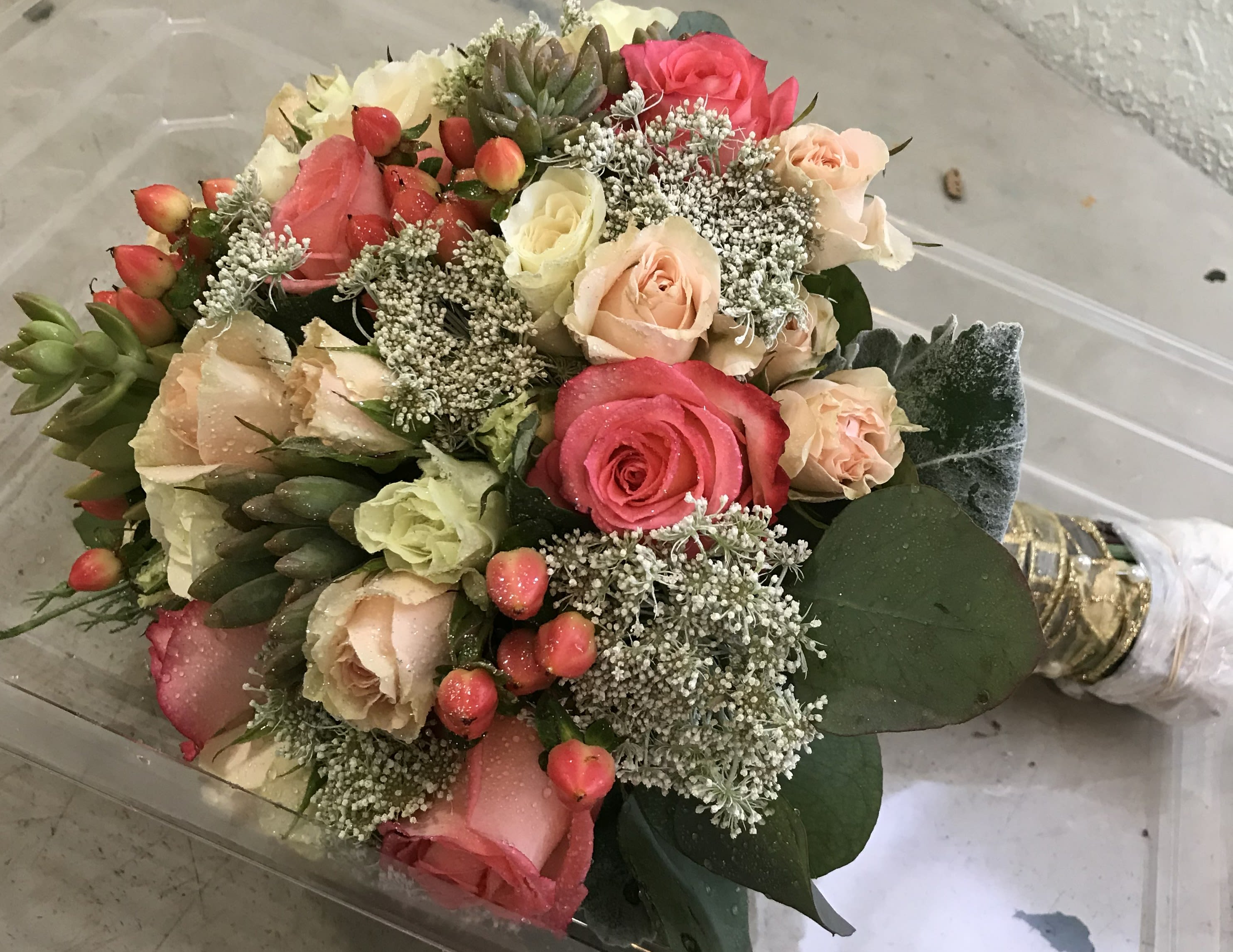 Exotic Wedding Bouquet  - A lush cluster of blush pink, peach, cream, and deeper pink roses creates a soft, romantic focal point-perfect as a wedding bouquet or an anniversary arrangement to make the person you're giving it to feel cherished. The pastel palette reads timeless and elegant, while the succulent-like accents and broad dark green leaves add surprising texture that keeps the look modern and garden-fresh.  This hand-tied flower arrangement features tightly clustered roses with small white filler blooms woven throughout for a light, airy touch. A bouquet wrap and decorative ribbon finish the stems for a polished presentation that's easy to carry. The mix of colors and textures ensures the bouquet photographs beautifully and complements a wide range of dresses and event styles.  Designed for ceremonies, vow renewals, or intimate celebrations, this bouquet sends a message of romance and thoughtful care-an especially meaningful way to mark an anniversary or to present at a wedding. Care: keep stems in water until just before use and change water daily to preserve freshness; sizing and stem counts are presented at checkout.