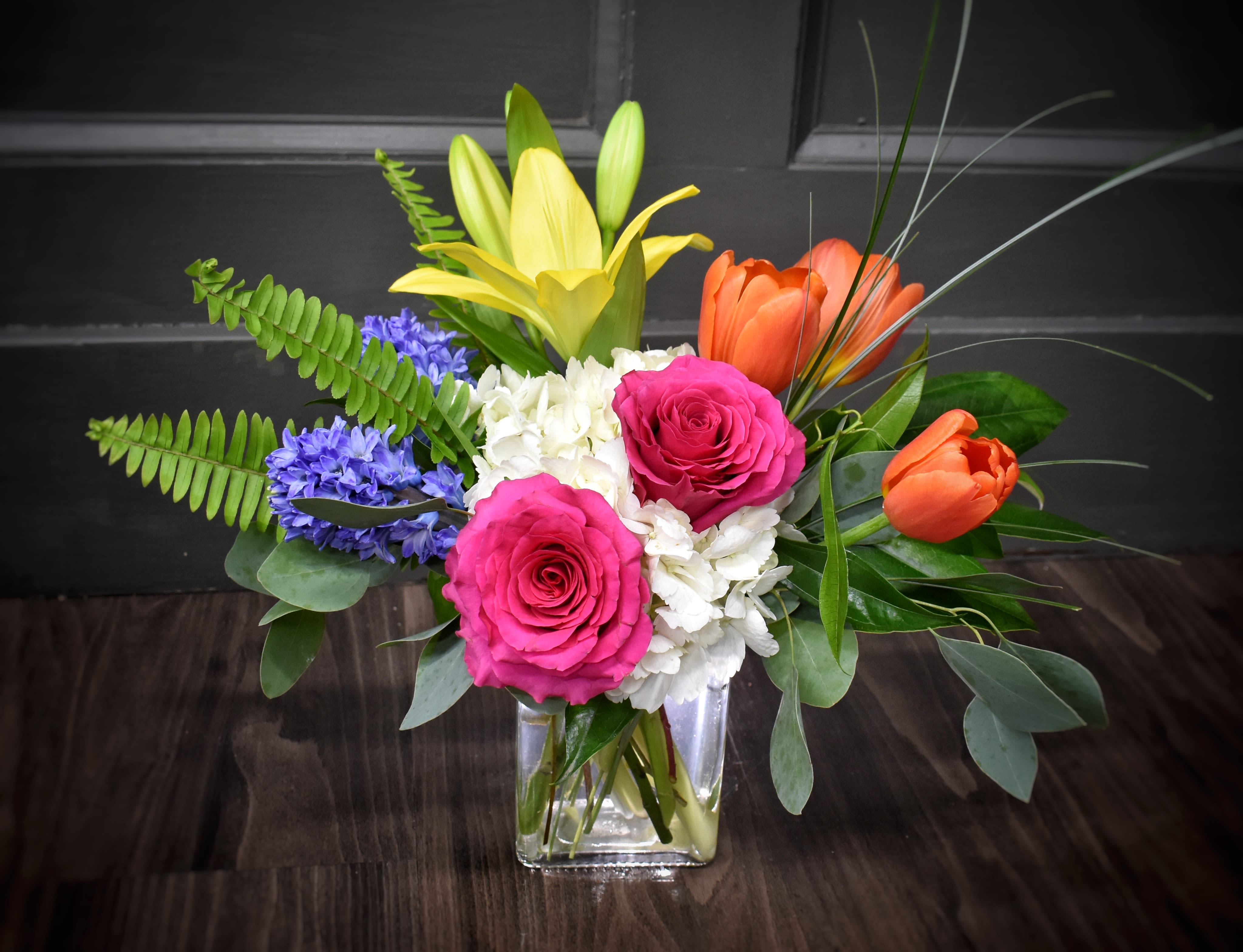 Meadow Fresh - Deluxe - Bright pink roses and sunlit yellow lilies meet orange tulips and purple accents in a lively, garden-fresh arrangement-an everyday mixed bouquet perfect for birthdays, congratulations, or a cheerful thank-you gift. This deluxe arrangement is presented in a clear square glass vase that keeps the focus on the saturated pinks, yellows, oranges, whites, and blue-purple accents. Blooms include roses, lilies, tulips, hyacinth and seasonal companions arranged full and loose for a natural, springtime look; stems extend outward for a dynamic presence on a table or desk. Sized to make an impression, it arrives ready to display-simply change the water every few days, trim stems as needed, and the bright colors will keep bringing smiles through the week.