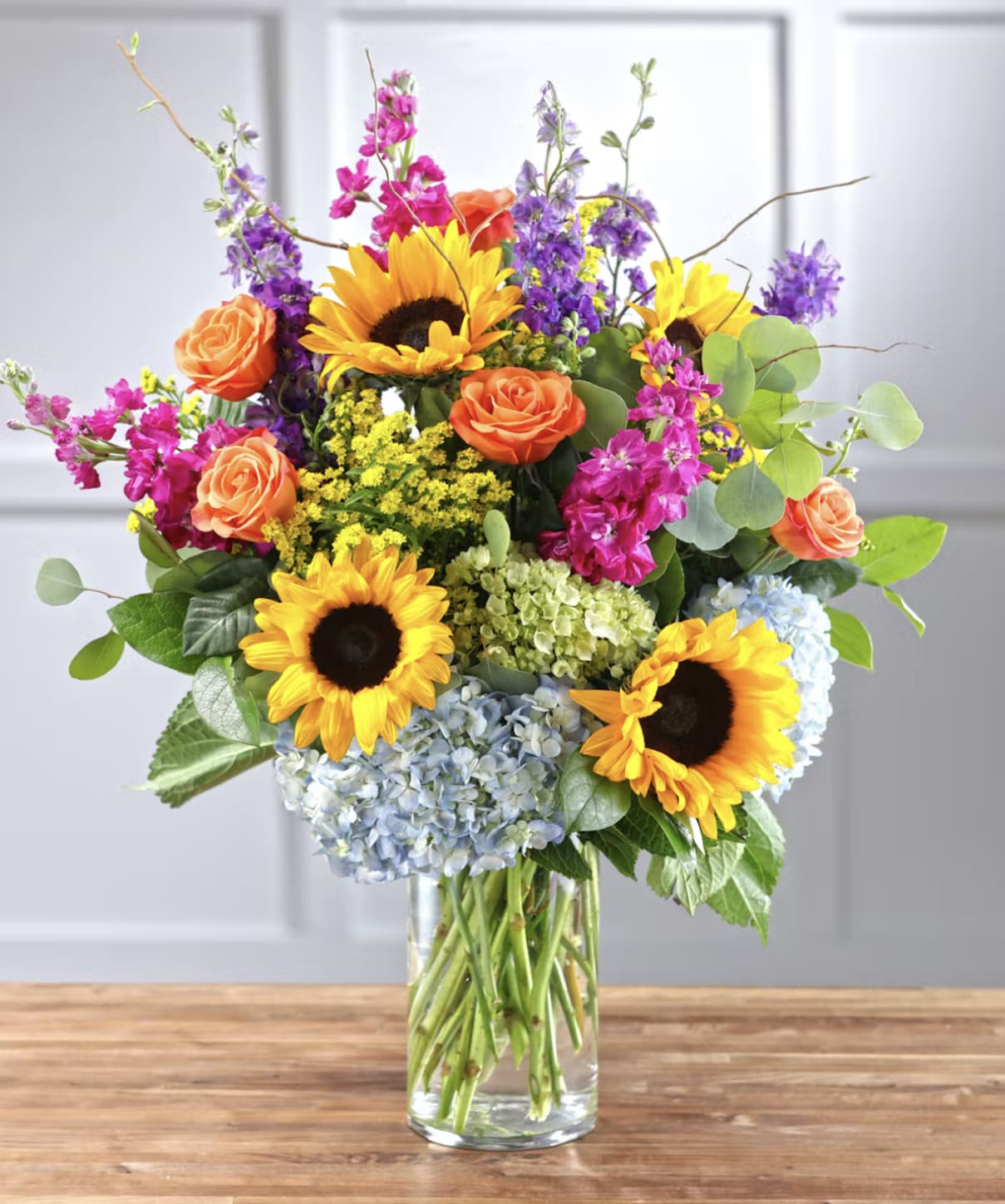 Mixed bouquet with sunflowers, roses, hydrangeas, and purple blooms in a glass vase