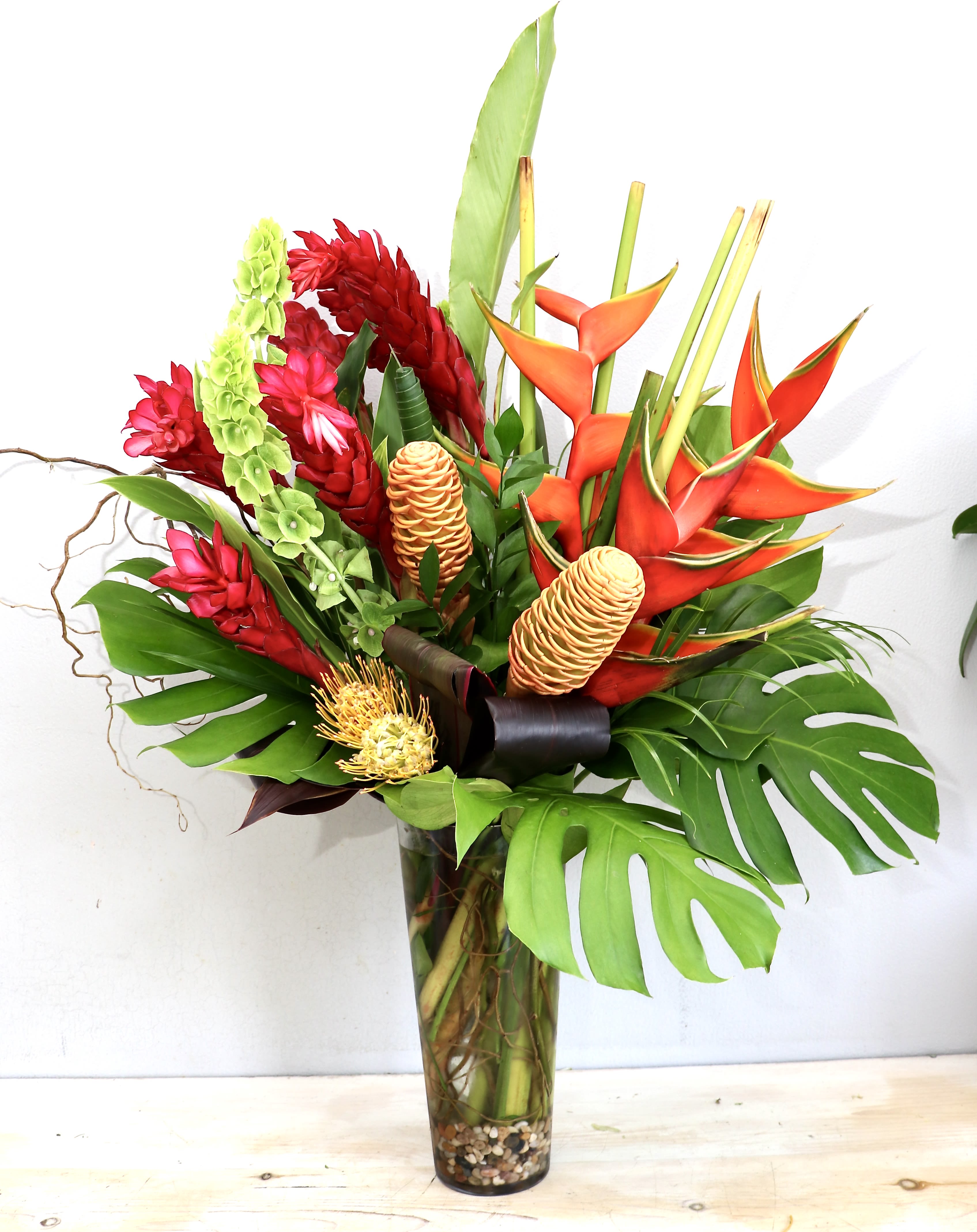 Tall Tropics - MGF - Bright orange heliconia-like blooms rise above glossy monstera-style leaves in this Tall Tropics flower arrangement, making a bold statement for a birthday bouquet or celebratory gift. The vertical silhouette and vivid mix of orange, red, pink, and green immediately read as energetic and exotic-the kind of arrangement that makes the person you're thinking of feel celebrated and noticed.  This tall tropical arrangement pairs clustered red flowers and a few soft pink accents with upright green stems for dramatic height and layered texture. It arrives in a clear glass vase with visible pebbles and roots, finished with a decorative ribbon for a polished presentation. The combination of sculptural foliage and bright tropical blooms creates a lush, gallery-worthy display that commands attention.  Perfect for anniversaries, milestone celebrations, or as a statement-get-well or congratulatory gift, Tall Tropics suits anyone who loves bold color and modern, architectural style. Because of its height and vertical design, place it where it can be seen from a distance-entryways, dining tables, or office lobbies-and enjoy the long-lasting visual impact of its tropical elements.