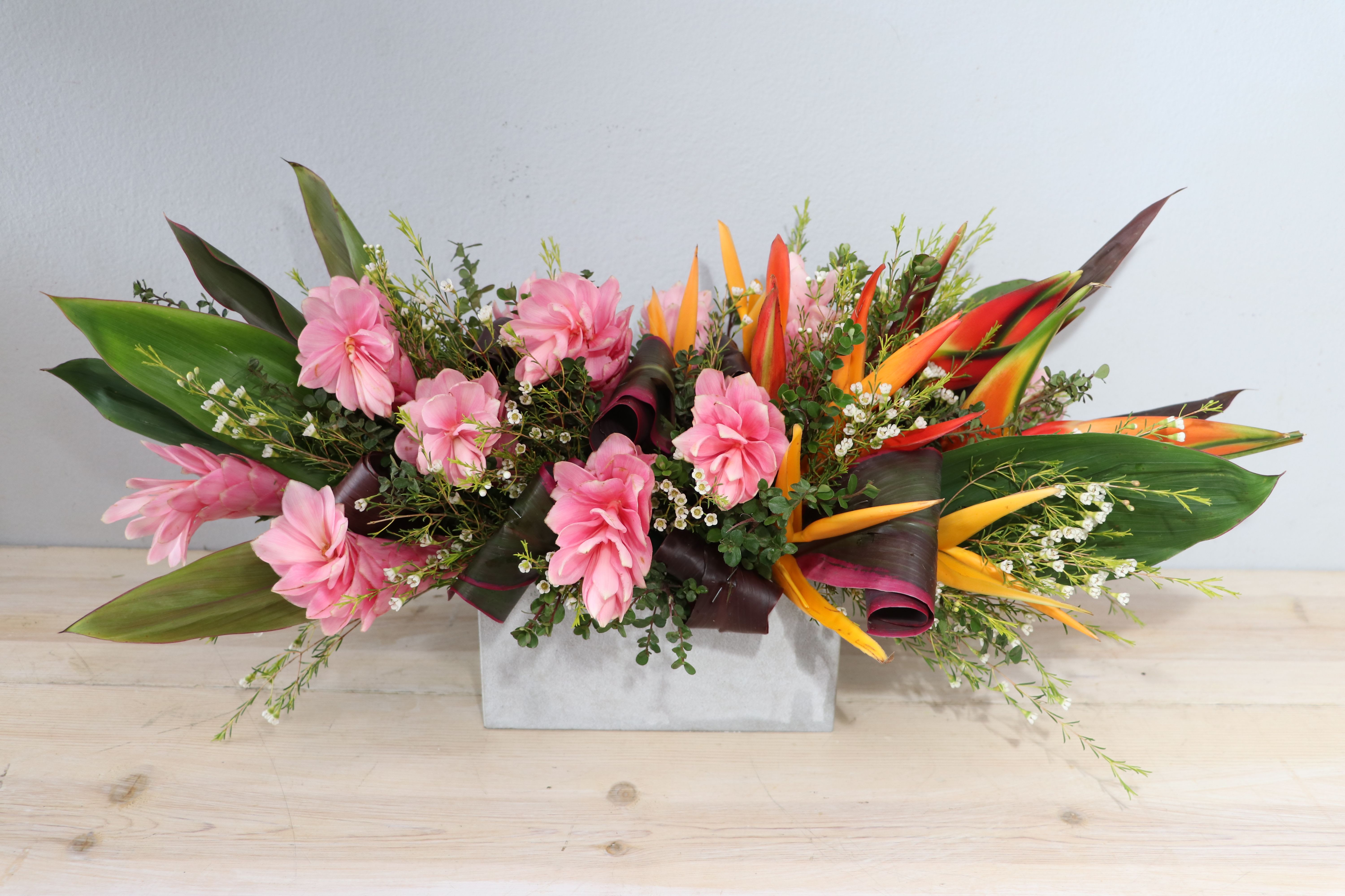 Ginger Tropics - MGF - Bright, celebratory and a little exotic - this Ginger Tropics flower arrangement is a vibrant choice for a birthday, congratulations, or a cheerful just-because gift. The low, wide composition and the rectangular box presentation give it a modern, easy-to-place silhouette that immediately reads as festive for the person you're thinking of.  Clusters of vivid orange and red tropical blooms, set against broad dark green leaves and delicate white filler flowers for contrast and texture. The compact design keeps everything low and lush, making it ideal for tabletops, desks, or as a centerpiece that won't block conversation.  Designed to make someone feel seen and celebrated, this arrangement delivers bold color and tropical energy without being overpowering. Care is simple-keep the floral foam hydrated and display in a cool spot away from direct sunlight to extend freshness.