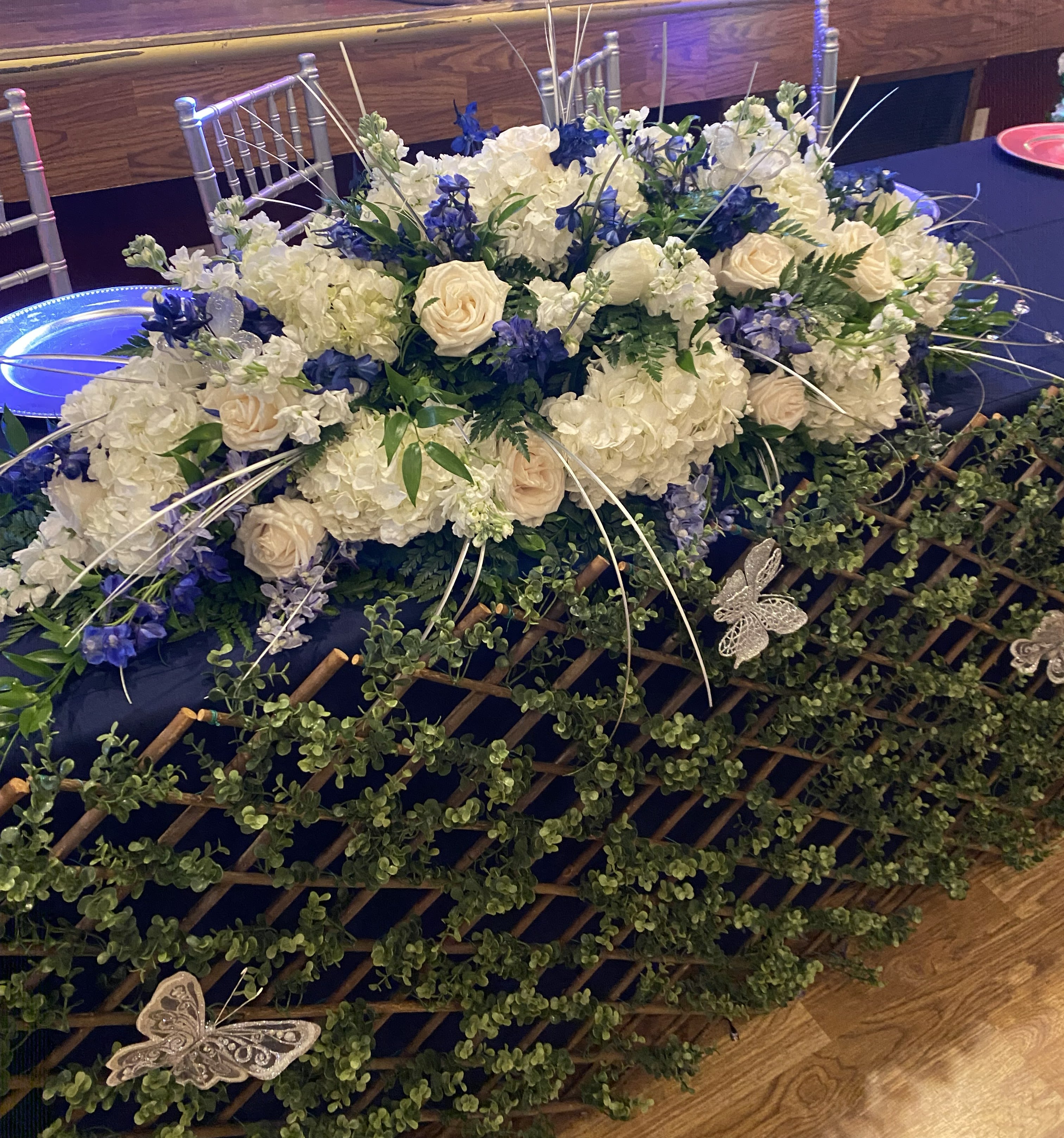 Main center piece for events - A elegant centerpiece that reads like a lush garden across your event table-this main center piece for events brings an elegant, wedding-ready look to any reception or formal gathering. The cool white-and-blue palette with touches of green and purple makes it ideal for weddings, anniversary dinners, or upscale showers where you want the honoree to feel truly celebrated; the arrangement's scale and sweeping silhouette also work beautifully for long banquet tables or head-table displays.  Dense clusters of white hydrangeas and cream roses form the soft, voluminous core, punctuated by vivid blue delphinium and other bright blue accents for depth and contrast. Trailing greenery cascades over a decorative lattice panel at the front of the table, while long white stems and white butterfly ornaments add a whimsical, refined finish. Designed to sit low and spread broadly, it complements standard banquet tables and pairs seamlessly with chairs and on-table decor without blocking sightlines.  Made to create a calm, sophisticated atmosphere, this centerpiece is the kind of floral that makes the person you're honoring feel seen and celebrated. Because it's crafted to cover and drape across the table, plan for a long table or clustered tables; light daily misting of the blooms and keeping the base in water will help the hydrangeas and roses stay fresh through the event.