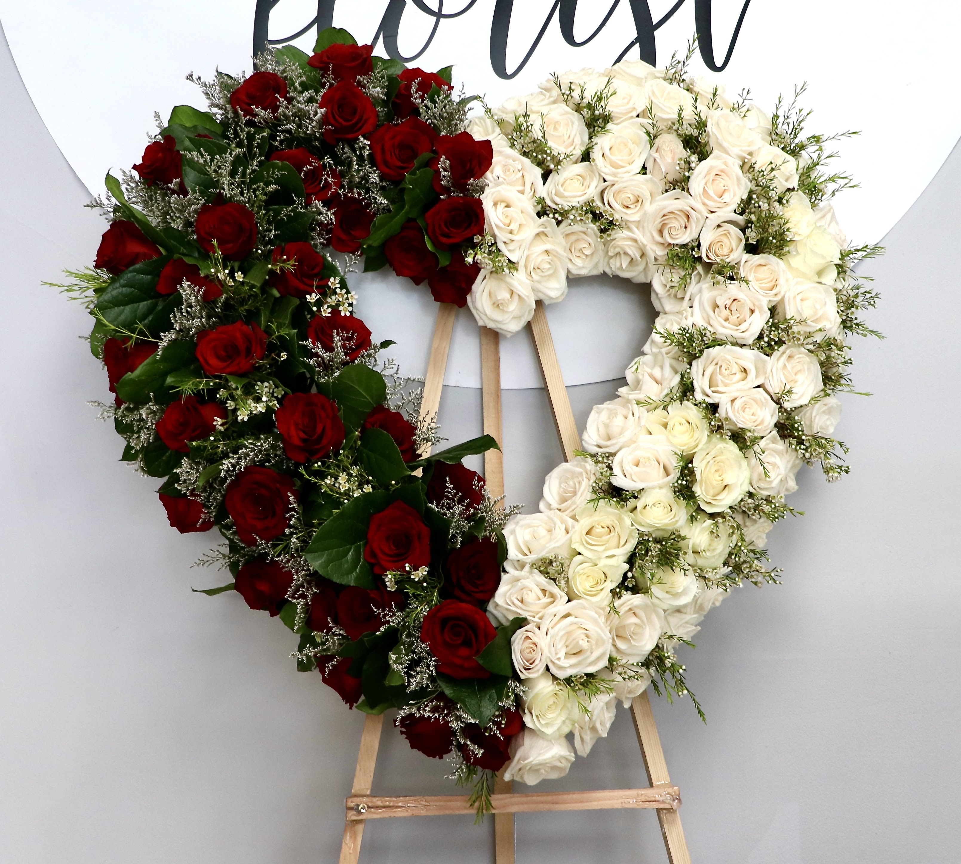 Red &amp; White Heart  - A striking heart-shaped sympathy arrangement made from densely clustered red and white roses creates an immediate visual and emotional impact-perfect for a memorial tribute or funeral floral display. The split of red roses on one side and white roses on the other offers a powerful contrast that reads as both romantic and reverent, making it an appropriate way to honor a loved one or mark a special remembrance.  Dark green foliage and small filler blooms are woven through the heart to add depth and a layered texture, giving the piece a full, sculpted appearance from every angle. Mounted on a sturdy wooden easel for formal presentation, this arrangement stands tall beside a casket or at a service to convey sympathy and devotion with clarity and grace.