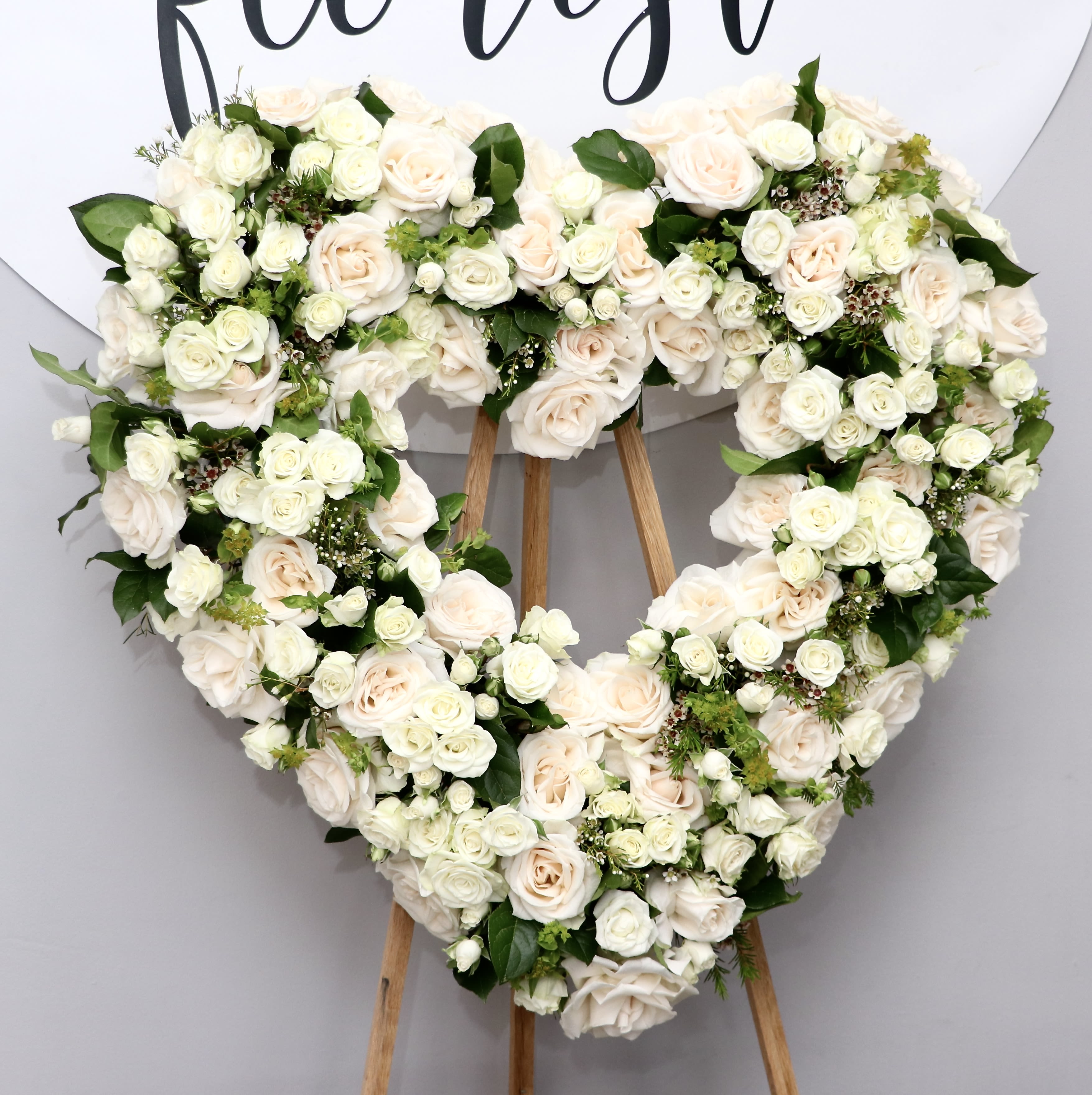 Garden Heart - A large heart-shaped funeral spray crafted from creamy white roses perfect as a sympathy arrangement to honor a beloved life. The Garden Heart makes a gentle, consoling statement-its densely packed roses form a plush, romantic silhouette that reads as reverent and heartfelt for memorial services, a funeral, or a keepsake tribute.  This arrangement combines full white roses with clusters of spray roses, the tight framing giving a lush, pillow-like texture. Dark green foliage is interspersed throughout to lift the colors and add contrast, and the piece is mounted on a sturdy wooden easel for prominent display at a service or graveside. The palette and shape convey tenderness and respect without being ornate.  Ideal for family members, close friends, or anyone you want to honor with a classic, sincere tribute, the Garden Heart communicates love and remembrance in a timeless way. Please note the arrangement is shipped ready to display on the included easel; water and fresh-care guidance is available on request to help preserve the blooms through services and viewings.