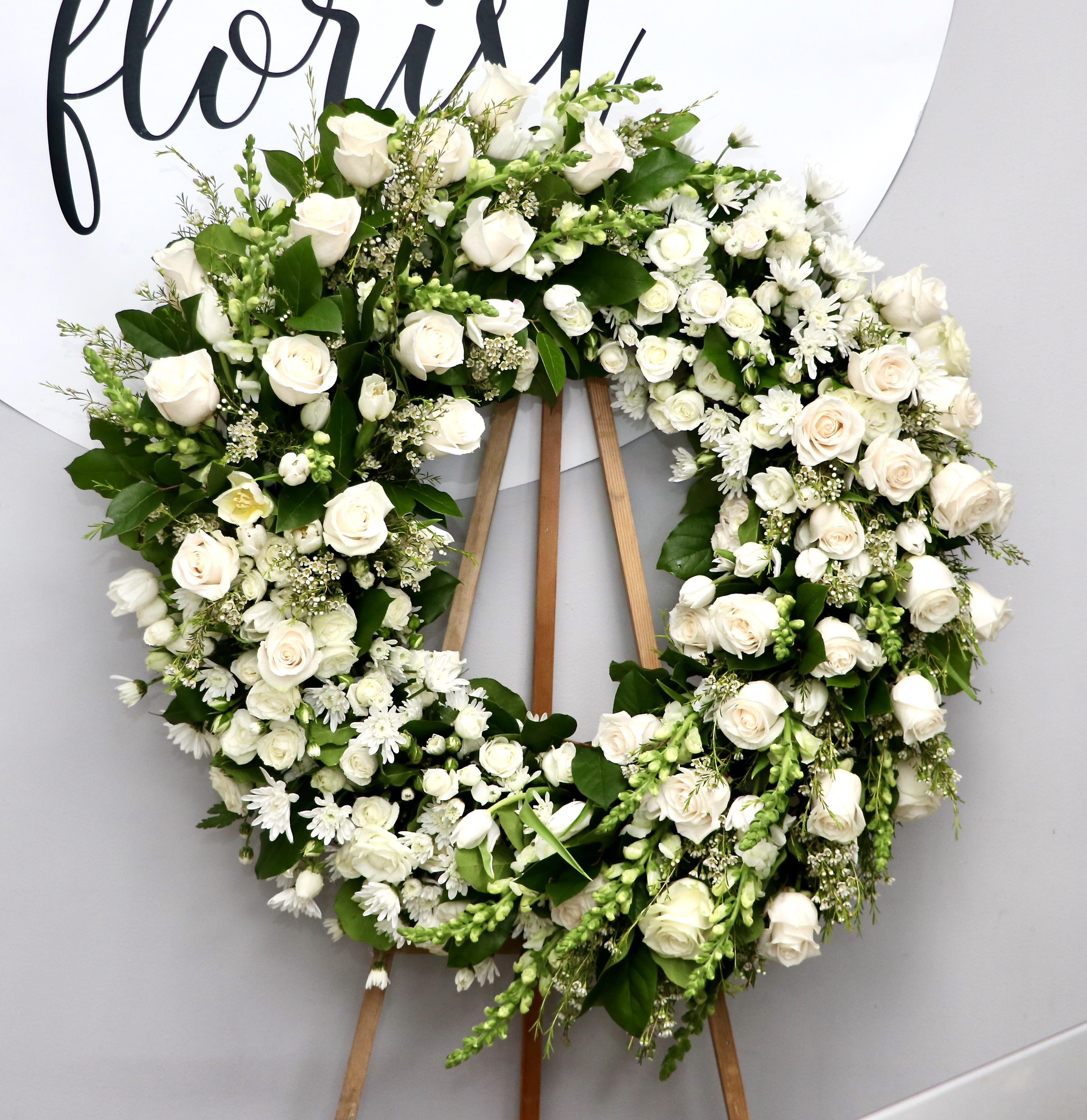 Elegant White Rose Sympathy Wreath - A classic circular sympathy wreath of pure white roses and daisy-like blooms offers a serene, comforting tribute for a memorial service or funeral. This flower arrangement and sympathy wreath is designed to express sympathy and respect; its clean white palette and formal wreath form convey reverence while keeping the focus on the person you're honoring.  The wreath is densely filled with white roses, small white daisy-style flowers and assorted fillers tucked into rich dark green foliage that gives crisp contrast and depth. Mounted on a sturdy wooden easel, the design reads as both elegant and subdued-large enough to make a respectful statement at a service while maintaining a tasteful, traditional silhouette.  Made to comfort the people receiving it, this arrangement is an appropriate way to say you're thinking of them and to honor a life. The wreath is delivered ready to display on the included easel; for best appearance, keep it away from direct heat and wind before the service.