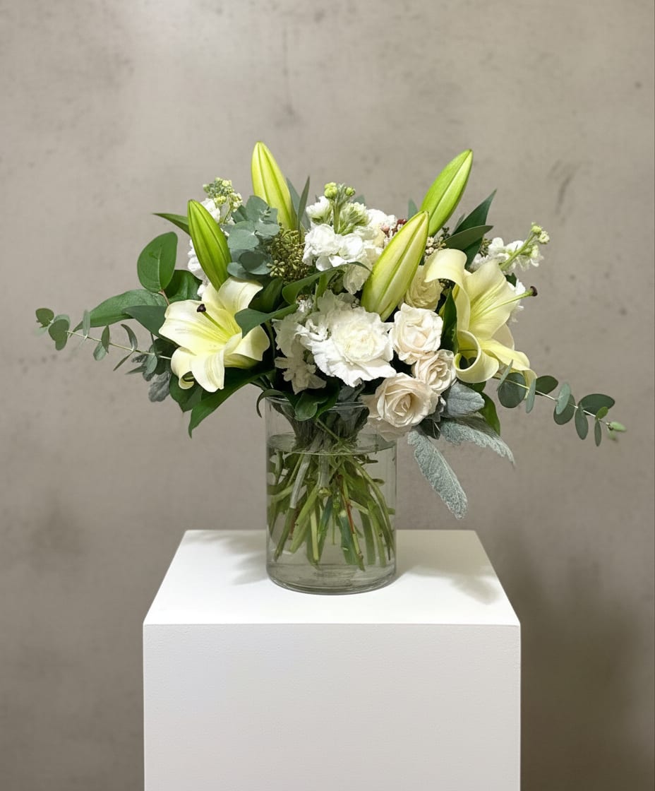 Cleopatra - Pale yellow lilies unfurl above a cloud of white roses and clustered blooms in an airy, garden-style bouquet-an elegant everyday arrangement perfect as a sympathy tribute, a thank-you gift, or a quiet celebration. The asymmetrical silhouette and soft color palette create a calming presence that makes the recipient feel gently seen and comforted.  White roses and creamy carnations mingle with clustered white flowers and eucalyptus, while broad green foliage reaches outward for a natural, just-picked look. Delivered in a clear glass vase, the design highlights stems and texture for a fresh, modern presentation with soft whites, fresh greens, and touches of yellow for warmth.  Choose this arrangement when you want to say you care without fuss-the kind of bouquet that brings quiet grace to a bedside, console table, or office. Standard sizing fits the pictured vase; water daily and trim stems every few days to keep lilies and roses looking their best.