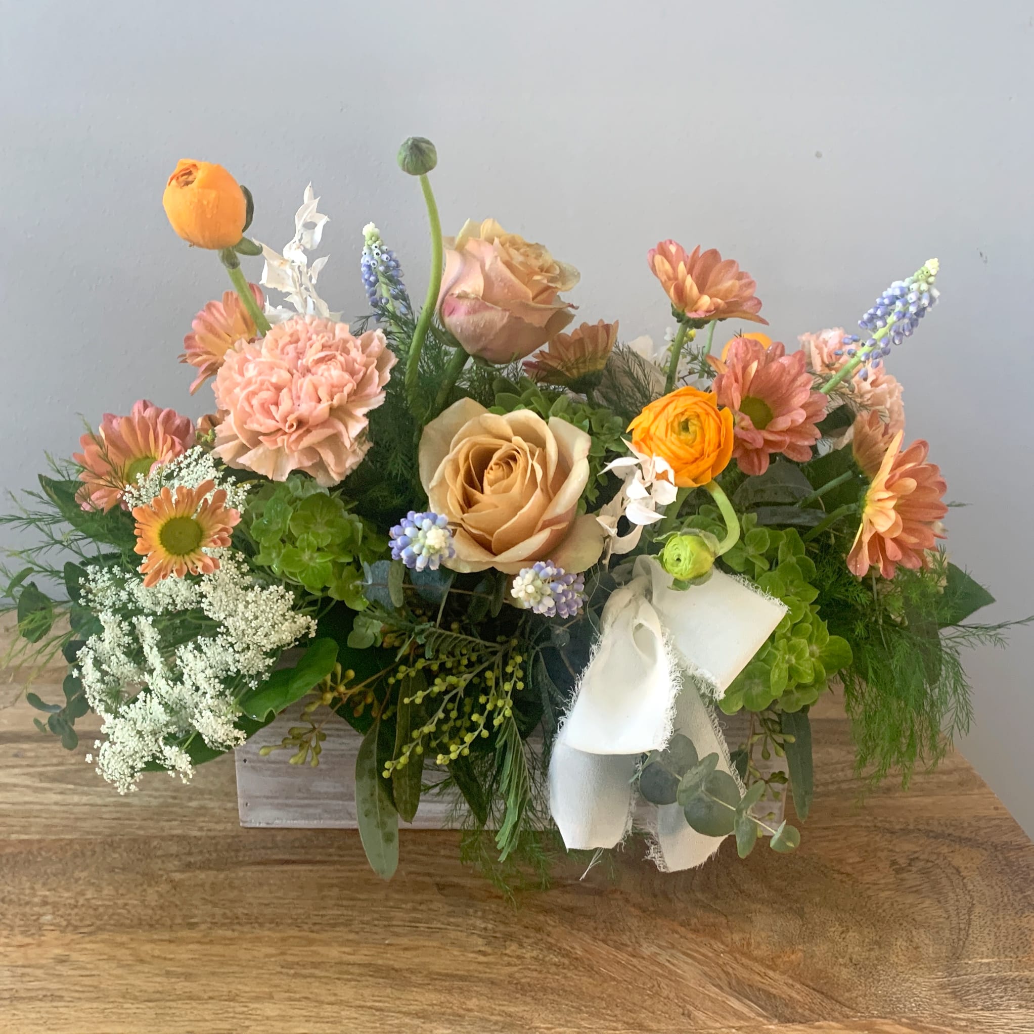 Low arrangement of peach roses, carnations, and orange blooms in a white wooden box with a fabric bow