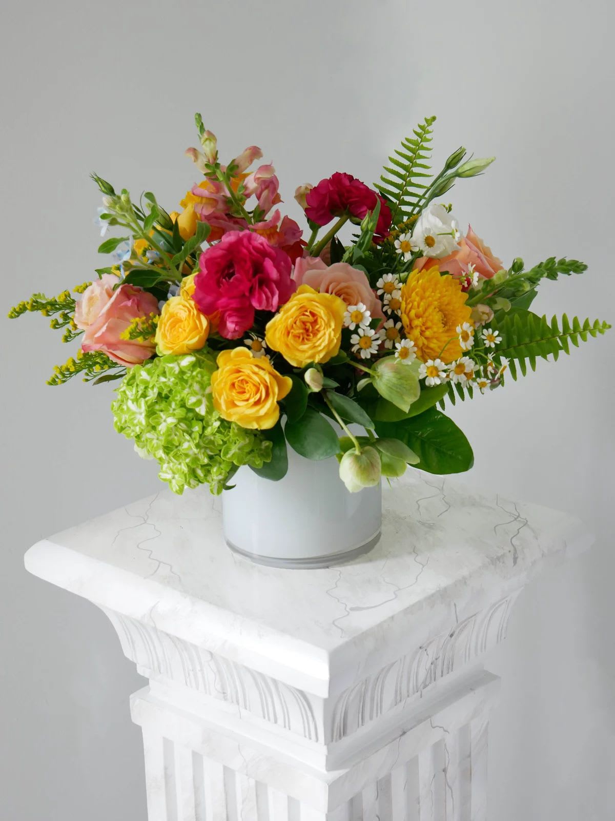 Garden Party Bliss - A cheerful garden-inspired arrangement bursting with vibrant seasonal blooms in shades of yellow, pink, peach, and green. Featuring roses, ranunculus, chamomile daisies, hydrangea, and fresh greenery, this playful and elegant design brings joy, sunshine, and natural beauty to any celebration or everyday moment.