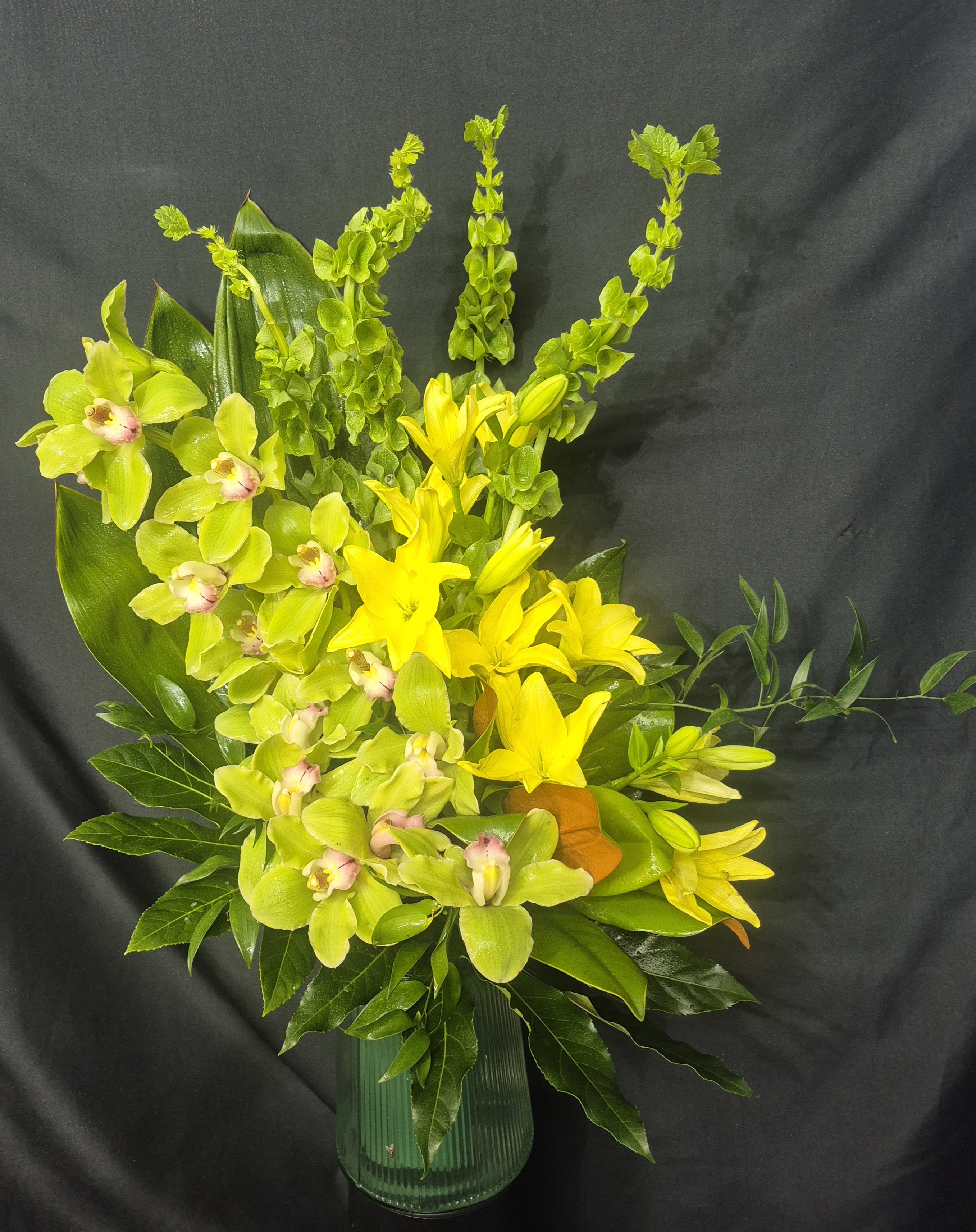Cymbidium Orchid Breeze - A vibrant and contemporary floral arrangement featuring elegant cymbidium orchids, radiant yellow lillies, and fresh seasonal greens. designed in a modern glass vase. This striking arrangement brings warmth, beauty, and sophistication to any celebration; birthday, anniversary, sympathy tribute, or special occasion. Orchid and lillie color can be changed upon request 