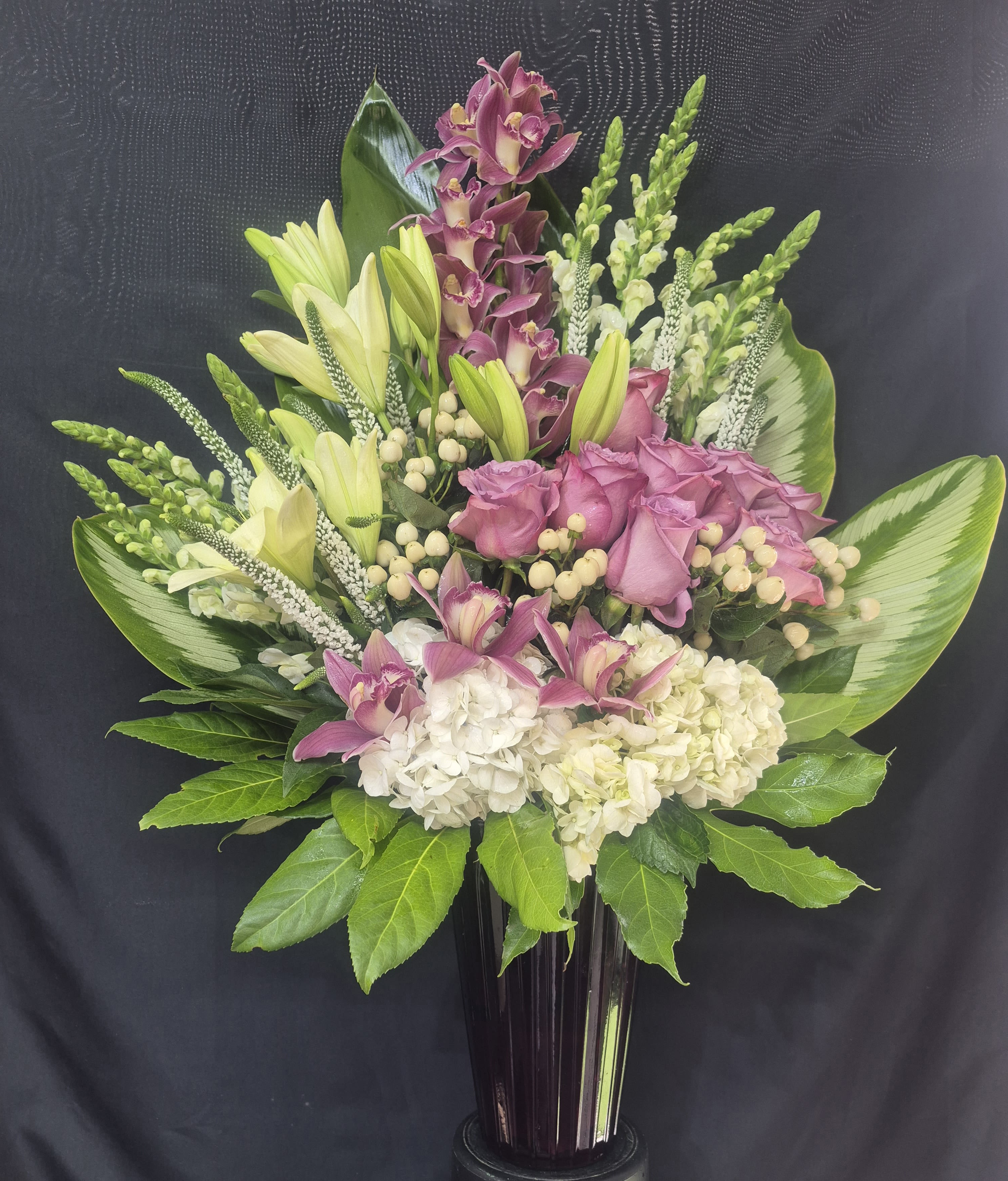 GRACEFUL ORCHID DREAMS - AN ELEGANT 39 INCHES TALL GARDEN INSPIRE ARRANGEMENT FEATURING LAVENDER ROSES, CYMBIDIUM ORCHIDS, WHITE HYDRANGEAS, LILLIES AND FRESH SEASONAL GREENERY. DESIGNED IN A TALL MODERN VASE, THIS LUXURIOUS FLORAL DISPLAY BLENDS SOFT PASTEL TONES WITH RICH TEXTURES, CREATING A GRACEFUL STATEMENT FOR CELEBRATIONS, SYMPATHY EXPRESSIONS, OR ANY SPECIAL OCCASIONS.