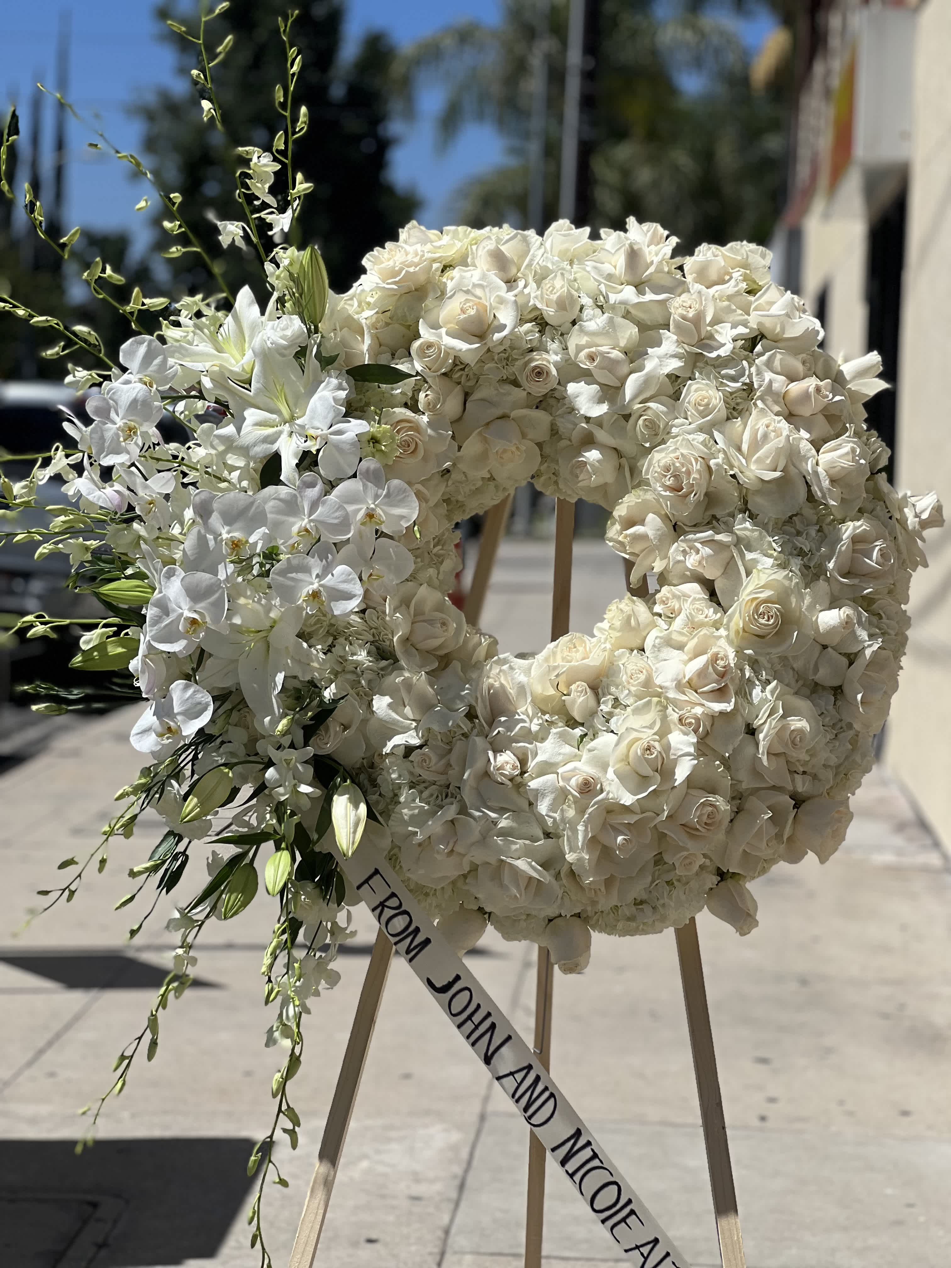 White floral wreath on an easel with a ribbon reading "FROM JOHN AND NICOLE"