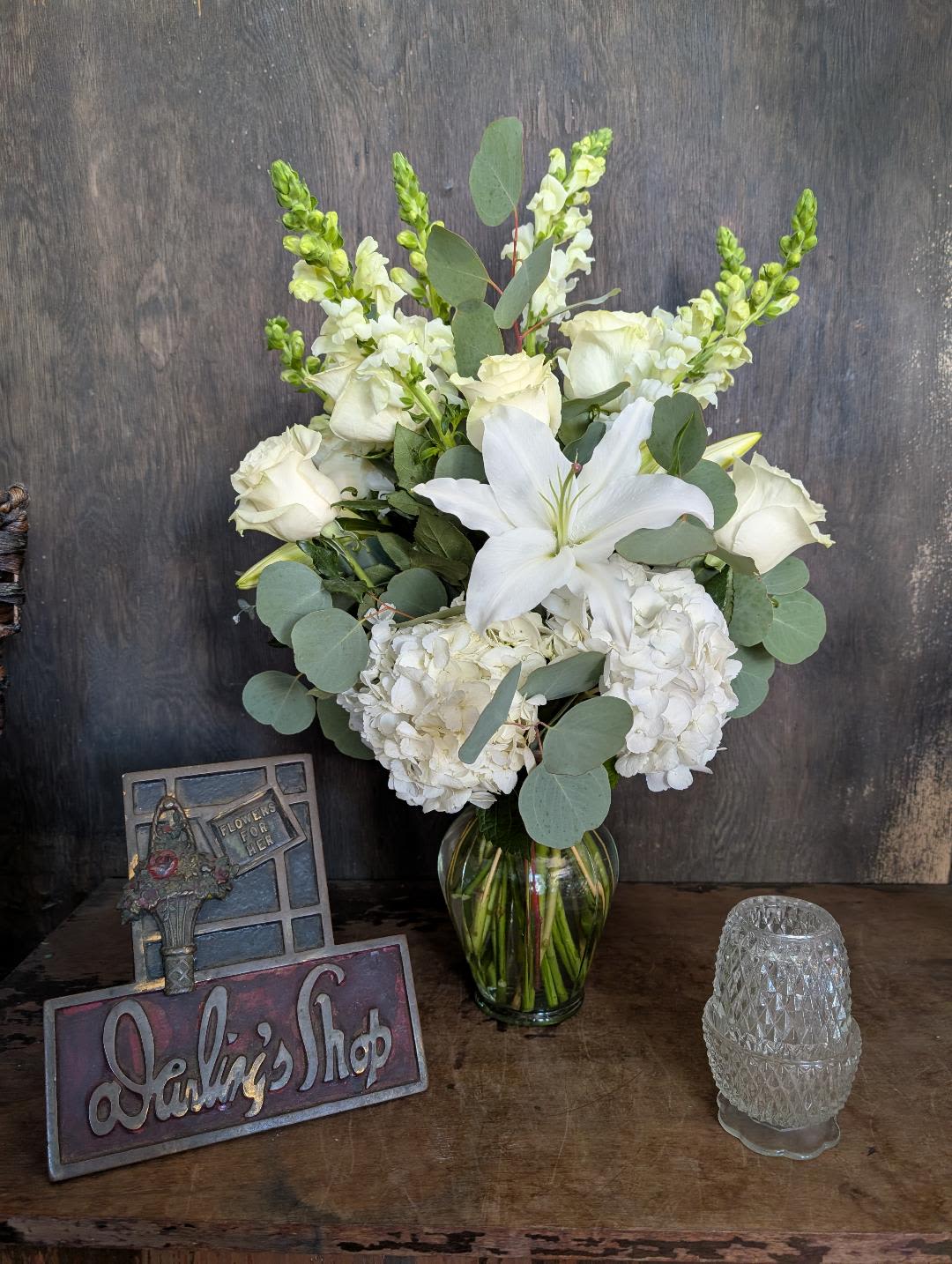 White floral arrangement in a glass vase with roses, lilies, and hydrangeas