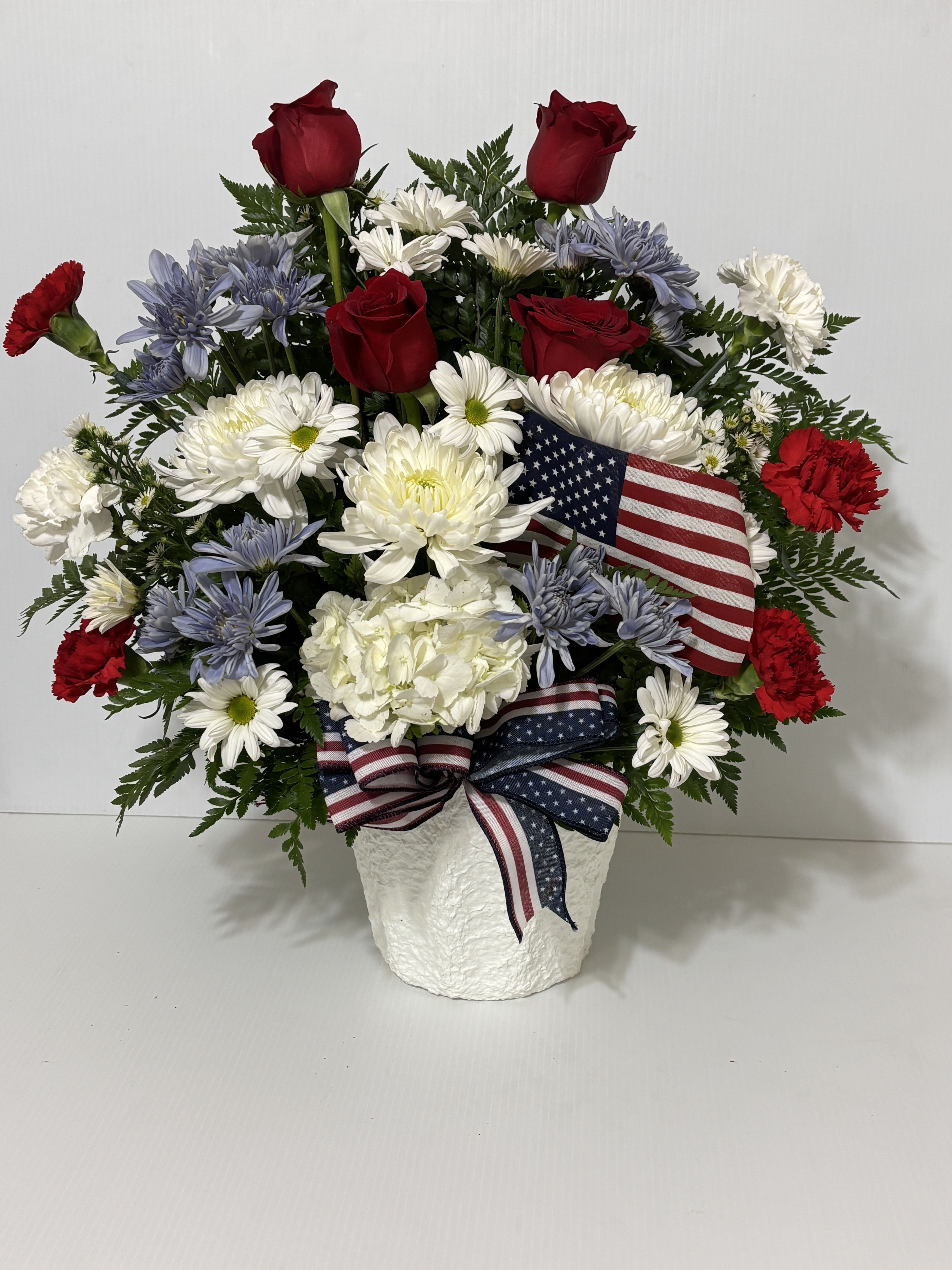 Red roses and white daisies in a white vase with an American flag ribbon