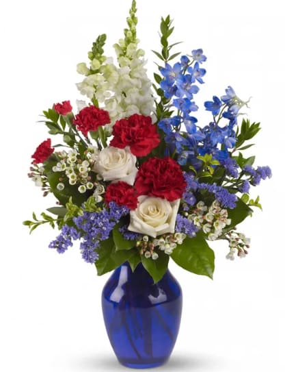 To Shining Sea - Show your patriotic spirit with this stunning floral arrangement, a perfect ode to national pride and unity. Featuring elegant White Roses symbolizing purity, Light Blue Delphinium representing tranquility, vibrant Red Carnations embodying strength, Red Miniature Carnations exuding passion, and pristine White Snapdragons adding a touch of grace. Adorned with lush floral greens, this arrangement is housed in a striking Cobalt Blue Vase, symbolizing loyalty and depth. 