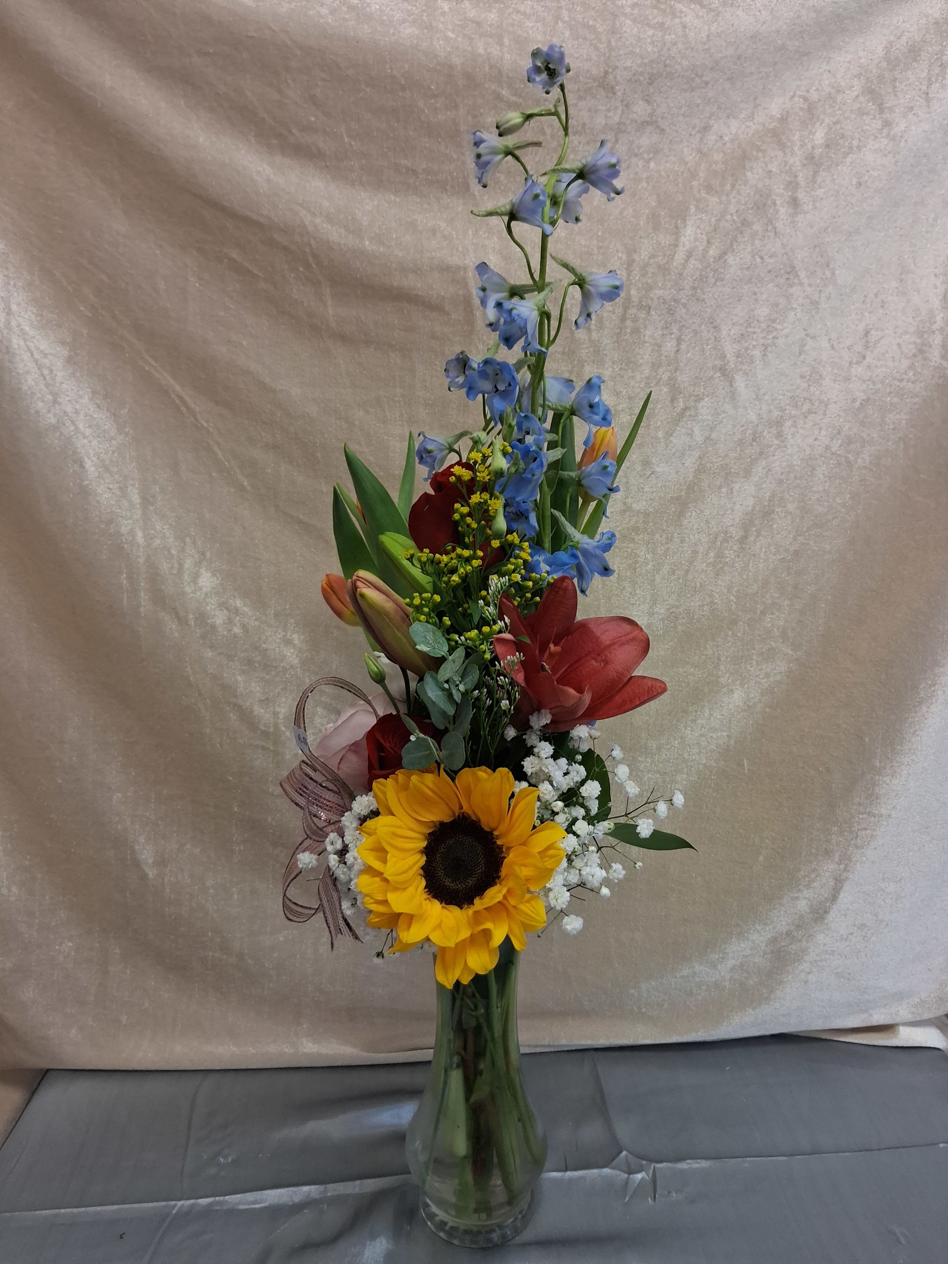 Get Well Arrangement  - A bright sunflower anchored at the base sets the tone for this Get Well arrangement-an uplifting sympathy-turned-cheerful bouquet designed to brighten a recovery room or bedside table. This flower arrangement for someone feeling under the weather pairs sunny yellow with vivid red lilies and cool blue delphinium, the color contrast crafted to feel both lively and reassuring for a get-well gift.  The composition is tall and vertical, with layered blooms and small white filler flowers that add airy texture as they rise above the focal sunflower. Delivered in a clear glass vase tied with a sheer decorative ribbon, the arrangement reads polished and gift-ready while still feeling warm and energetic. The mix of sunflowers, delphinium, lilies, and delicate fillers creates visual interest and a pleasing balance of bold color and soft accents.  Perfect for a friend, colleague, or family member recovering from illness or surgery, this bouquet sends a message of encouragement and sunshine. The clear vase holds the stems neatly for easy display; standard care-fresh water and a cool spot away from direct sun-will help the blooms last and keep the recipient smiling.