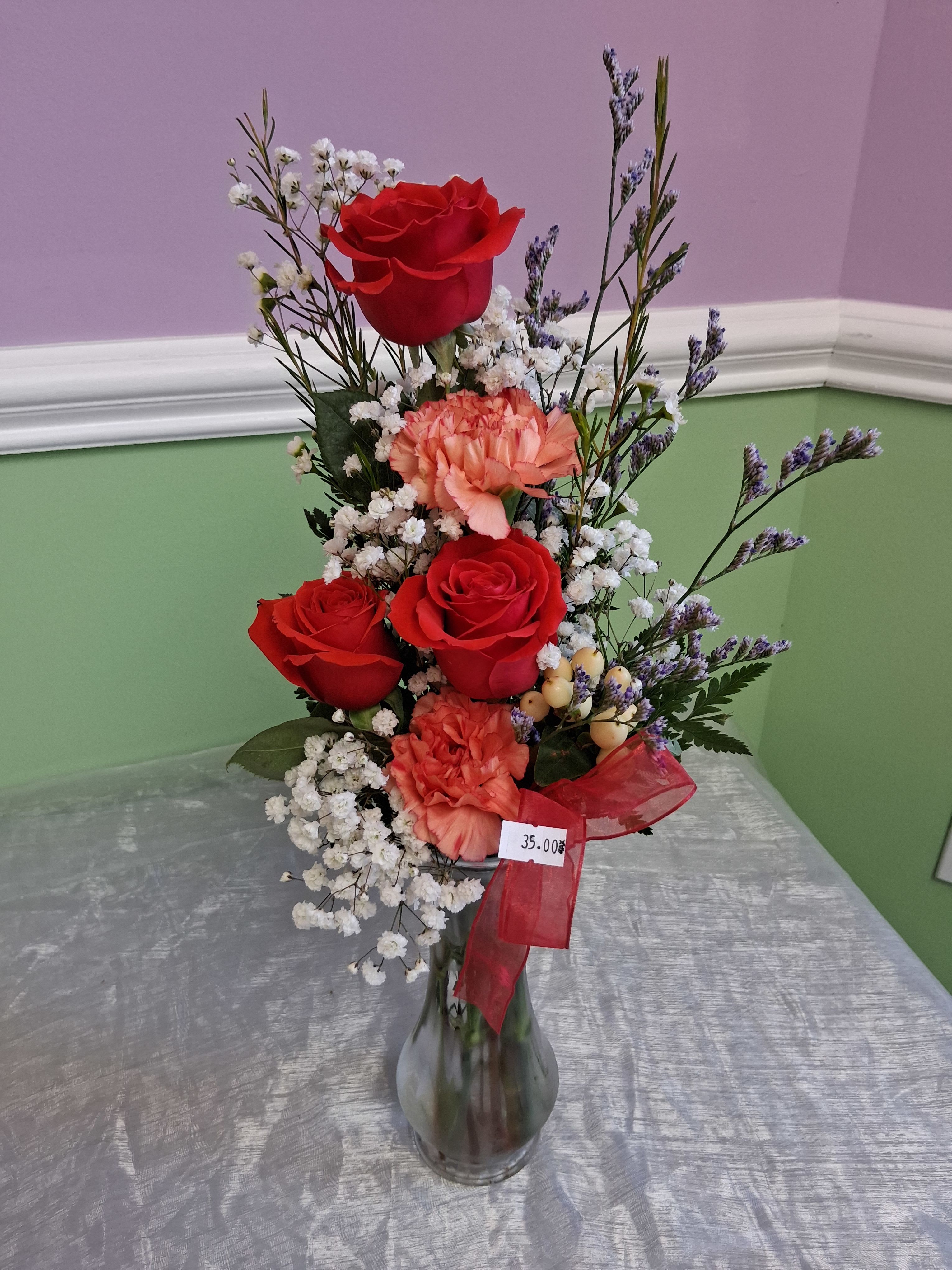 3 Roses with Carnation - Three romantic red roses take center stage in this petite flower arrangement, perfect as an anniversary roses bouquet or a Valentine's Day gift that feels both intimate and cheerful. The mix reads like an everyday romantic surprise for the person you're thinking of-she'll feel noticed and cherished by the classic red roses paired with soft-textured coordinating blooms.  Alongside the three red roses are peach carnations, airy white baby's breath, and small purple filler stems that add height and a delicate contrast to the fuller blooms. The stems come arranged in a clear glass vase tied with a red ribbon; a visible price tag gives it a casual, shop-display charm. This compact arrangement suits tabletops and desks-order for next-day delivery or pickup to make a simple, heartfelt moment feel special.